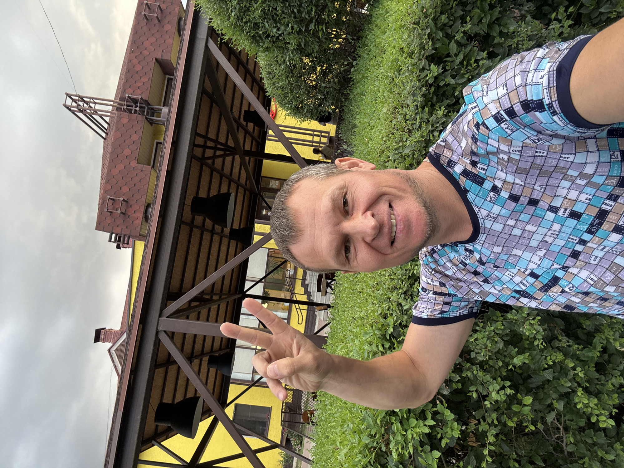 A middle-aged man with short, light brown hair and a light stubble is smiling and making a peace sign with his right hand. He is wearing a short-sleeved t-shirt with a checkered pattern of blue, grey, and white squares. He is standing in front of a dense green hedge. Behind the hedge, a yellow building with a brown, angled roof structure is visible. Several large, black pendant lamps hang from the roof structure. The sky above is overcast and grey. The building appears to be a restaurant or cafe. This photo was taken in Oskemen, Kazakhstan.