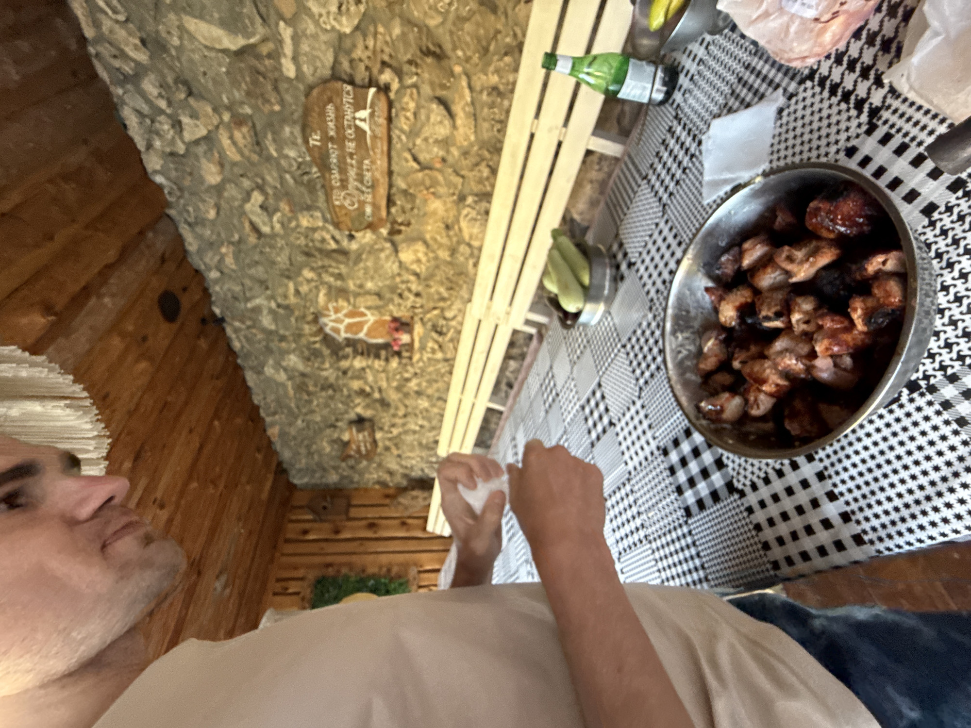 A man is standing next to a table covered with a black and white checkered tablecloth. He is wearing a light-colored t-shirt and jeans. His hands are occupied with what appears to be a white napkin. On the table, a metal bowl is filled with cooked meat pieces, likely shashlik. There are also several green vegetables, possibly zucchini, in a smaller metal bowl. A green glass bottle, a roll of paper towels, and a plastic bag are also visible on the table. In the background, a stone wall is adorned with a wooden plaque containing text in Cyrillic. Behind the table, a wooden bench is placed against the wall. The ceiling is made of wooden planks, and a decorative element hangs from it. The overall setting appears to be an outdoor or semi-outdoor dining area. The location is specified as Krasnomakskoye rural settlement, Ukraine.