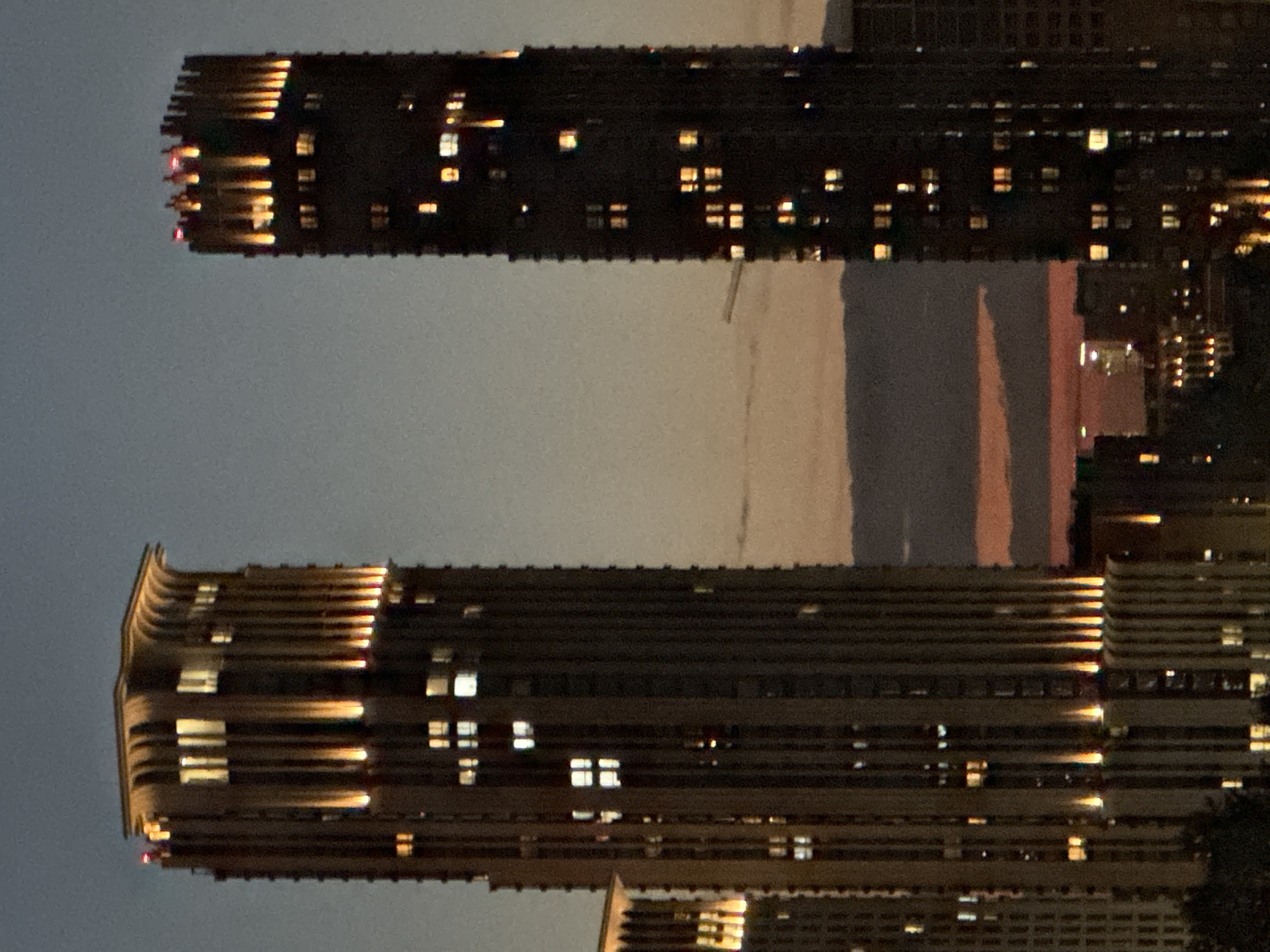 This media file presents an urban cityscape at night, likely viewed from a height in Moscow, Russia. Two prominent, tall modern high-rise buildings dominate the frame.
The building on the left side (when viewed upright, as the provided image is rotated) features a distinct, subtly curved architectural design at its upper sections. Horizontal ledges or balconies line its facade, reflecting warm light from numerous internal windows. These windows glow with scattered light, creating a mosaic of bright rectangles against the building's dark exterior.
The building on the right is similar in height and contemporary design, also exhibiting a dark facade punctuated by numerous illuminated windows. Its upper portion also shows architectural details that catch and reflect light.
Between and behind these two primary structures, the dark sky transitions to a softer, warmer glow near the horizon, displaying subtle orange and reddish hues, indicative of a lingering sunset or the distant luminescence of city lights. Other less defined buildings with faint lights are visible further in the background.
The scene captures the quiet, illuminated presence of urban architecture after dusk. No people or specific activities are visible, and there is no discernible text. The image conveys a calm, observational perspective of a metropolitan area at night.