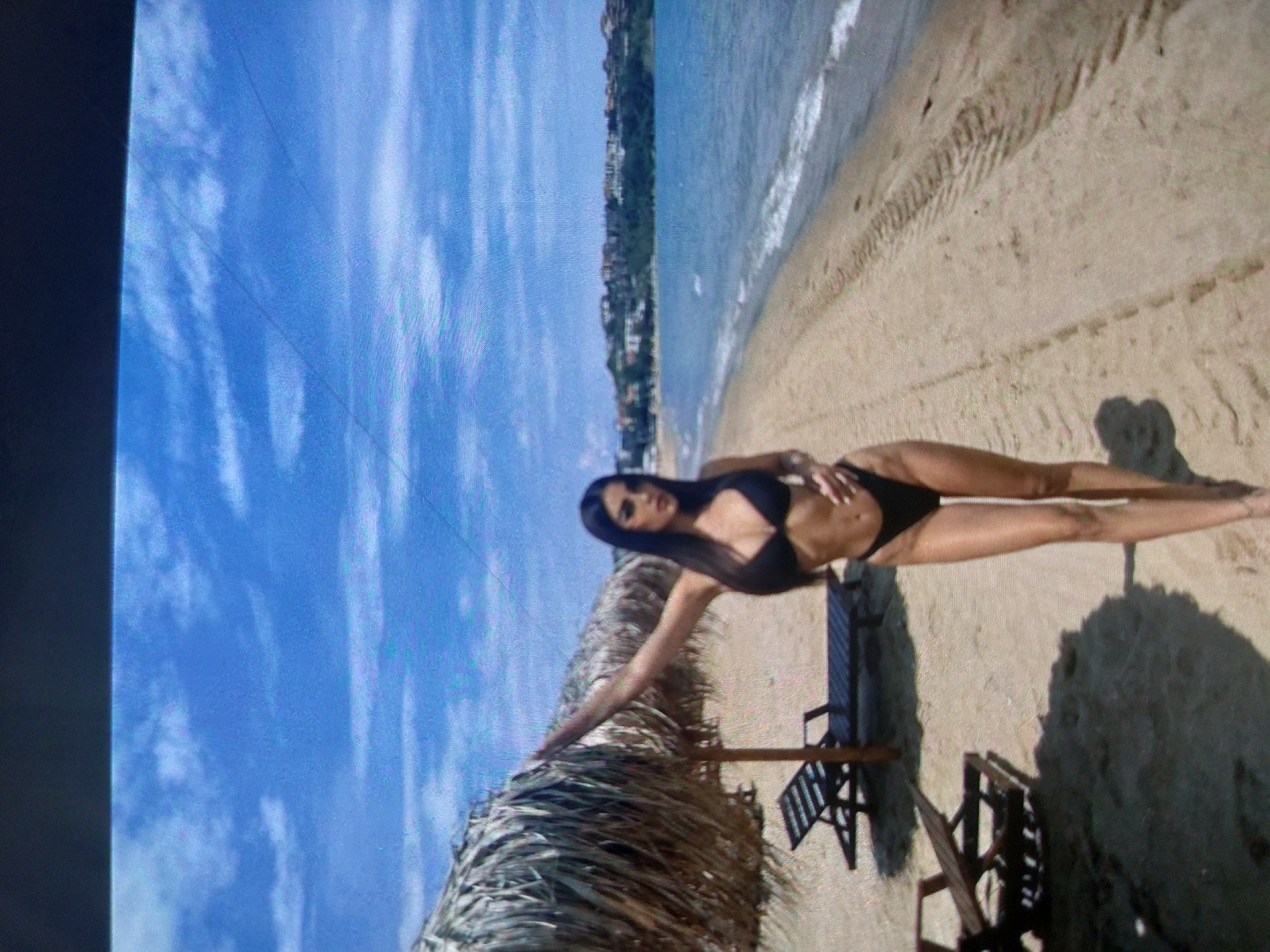 The image depicts a woman lying on a sandy beach in Tsarevo, Bulgaria, based on the appearance of the coastline and architecture in the background. The woman is wearing a black bikini and has dark hair. She is positioned near a sun umbrella made of straw and several wooden beach chairs. The sky is blue with scattered clouds, and the sea is visible in the background. The beach has tire tracks or marks on the sand.