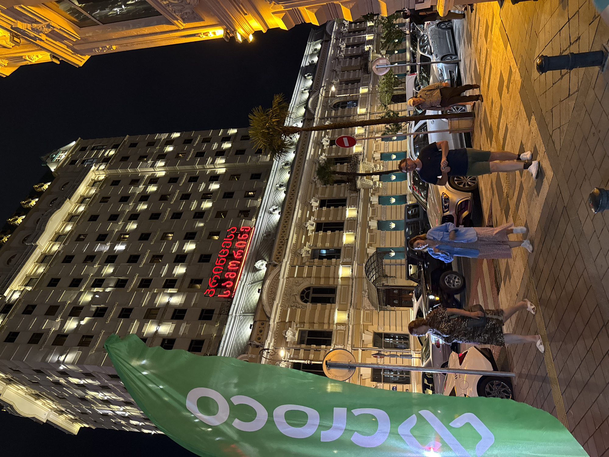 This is a street-level, wide-angle night shot of a well-lit building with a large red sign in Georgian script reading "კლასიკა" which translates to "Classica". The building is tall with numerous windows and is illuminated with warm, golden lights. In the foreground, a large green banner with white text reading "croco" is visible, partially obscuring the building. Several people are walking on the sidewalk in front of the building, and cars are parked and driving on the street. The scene appears to be a busy urban environment, possibly a hotel or a commercial establishment. The architecture suggests a historic European city. The presence of palm trees and the style of the building's facade are characteristic of Batumi, Georgia.