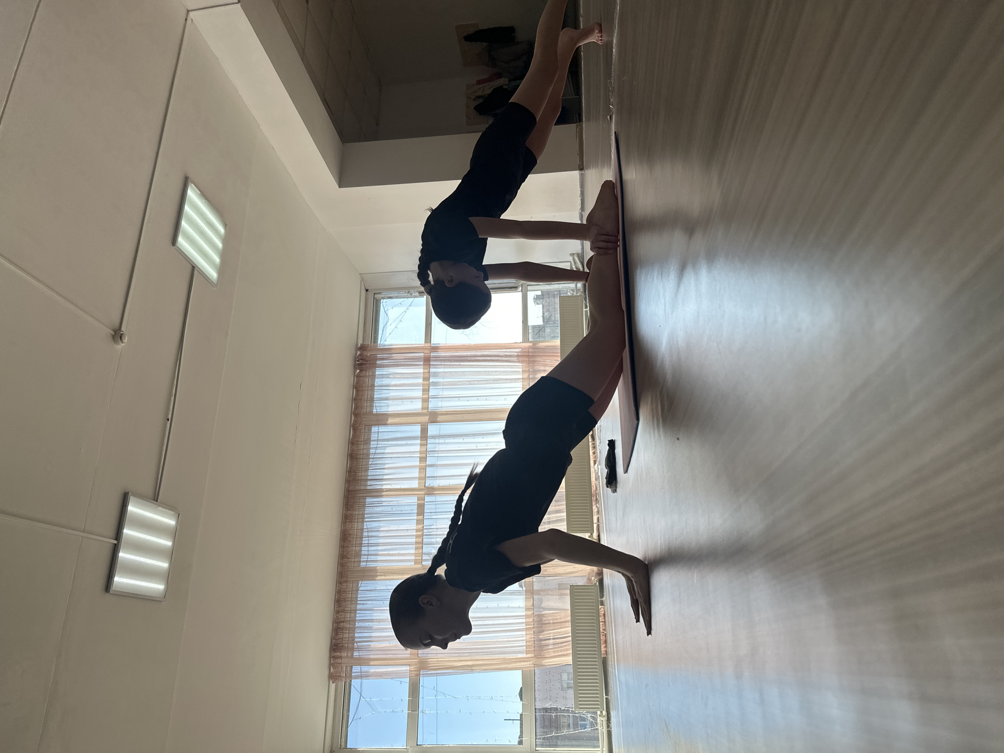 The image depicts an indoor exercise room in Votkinsk, Russia. Two individuals are performing exercises on a light brown, reflective floor.
The individual on the left, positioned towards the foreground, is on a pink yoga mat. She is propped on her hands and knees with her torso elevated, in a pose resembling a modified push-up or upward-facing dog. She wears a black t-shirt and black shorts, and her dark hair is styled in a single braid. A dark fabric item is visible on the mat near her knees.
The individual on the right is in a plank position, facing away from the camera, supported by hands and bare feet. She also wears a black t-shirt and black shorts. Part of a dark-colored yoga mat is visible beneath her feet.
The room features a wall of large windows on the left, covered by translucent light brown sheer curtains. Through the windows, a clear blue sky is visible above an urban landscape of buildings. Decorative string lights are strung horizontally across the window panes. White radiators are installed along the wall beneath the windows. The ceiling is white, with exposed conduit running across it, and features two rectangular overhead light fixtures, each containing four illuminated linear bulbs. A small, round, white object, possibly a smoke detector or sensor, is mounted on the ceiling. White walls define the space, with a doorway or opening on the right leading into a dimly lit area where clothing items hang on a rack or hooks. The room is illuminated by both natural light from the windows and artificial light from the ceiling fixtures.