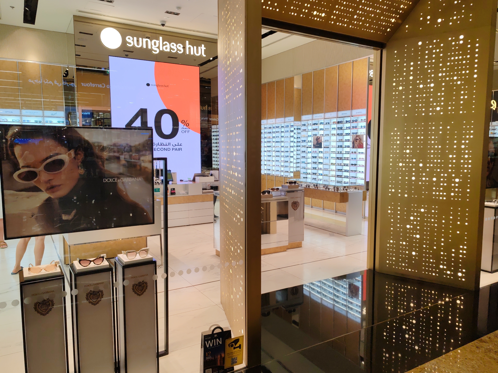 The image captures the exterior view of a Sunglass Hut store, likely located within a shopping mall in Dubai, United Arab Emirates. The store features a large illuminated sign that reads "sunglass hut" and a digital display showcasing a "40% OFF" promotion for the "SECOND PAIR" of sunglasses, with text in Arabic also present.
In the foreground, a display showcases three pairs of Dolce & Gabbana sunglasses on individual pedestals. The pedestals are branded with the Dolce & Gabbana logo. Adjacent to the display, on the floor, are two promotional signs; one with the word "WIN" and another displaying a CCTV camera icon.
The interior of the store is visible through an open entrance, revealing rows of sunglasses displayed on shelves and on a wall. The store's interior is modern and well-lit, with warm-toned cabinetry and sleek displays. The overall environment appears to be a high-end retail setting, common in Dubai's luxury shopping destinations.