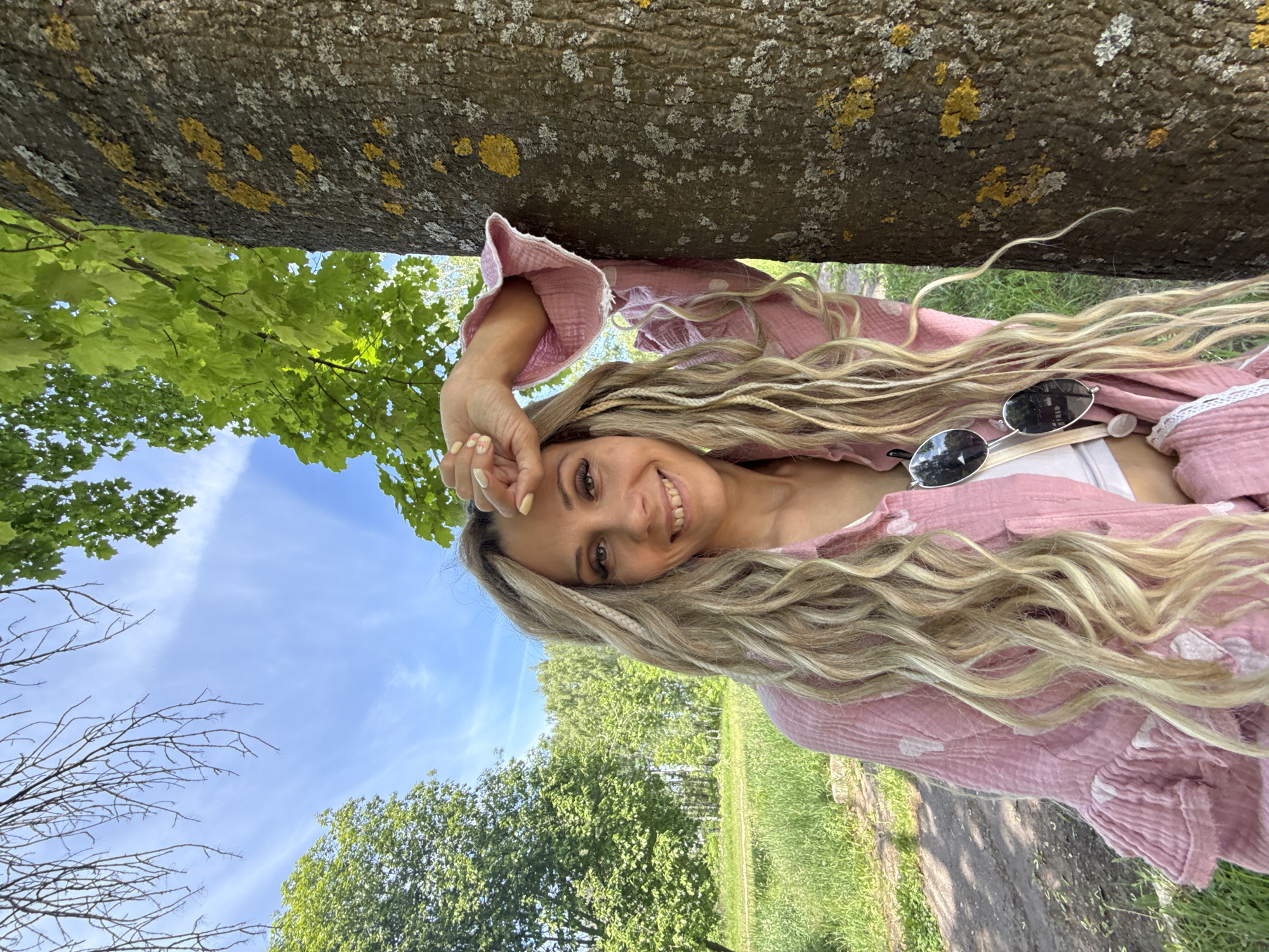 This image depicts a smiling woman posing outdoors in what is described as Rusanovo, Russia. The woman, appearing to be in her 20s or 30s, has long, wavy blonde hair with several lighter, braided sections. She wears a light pink, textured long-sleeved shirt, possibly made of gauze, with subtle white polka dots and ruffled cuffs. A white top is visible underneath, and dark sunglasses hang from her neckline. Her left hand, adorned with light yellow nail polish, is raised to her forehead, partially shielding her eyes from the sun. She looks directly at the viewer with a friendly smile.
She is leaning against the trunk of a large tree on the right side of the frame. The tree trunk is dark-barked and covered with patches of white and bright yellow lichen. The background reveals a bright blue sky with wispy white clouds. Lush green trees and grass fill the mid-ground, suggesting a park or wooded area. A dirt path is visible winding through the grass on the left side of the background. Some bare tree branches are also visible against the sky in the upper left corner. The scene is brightly lit by natural sunlight, indicating a clear day.