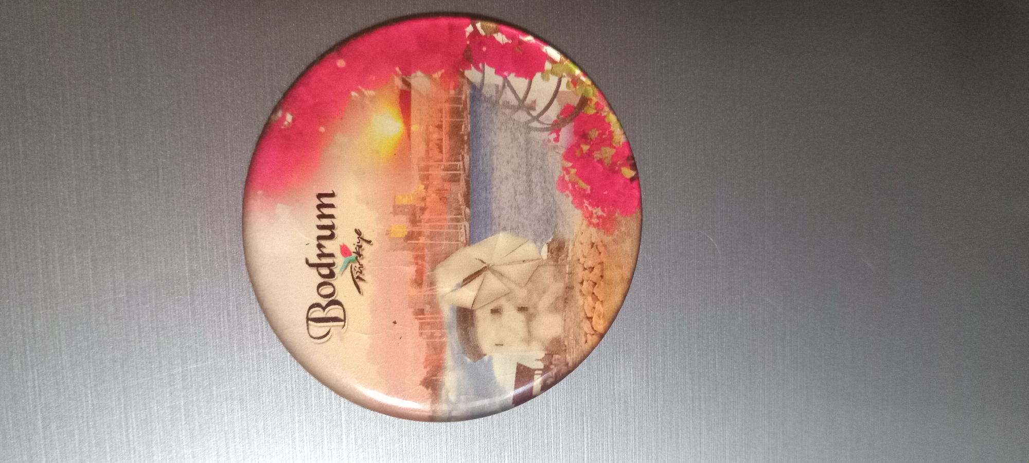 This is an image of a round magnet with a picture on it, placed on a grey, textured surface, likely a refrigerator or metal cabinet. The magnet depicts a scene of a seaside town with bougainvillea flowers.
In the foreground of the magnet's image, text reads "Bodrum" in a large, stylized font. Below this, smaller text says "Türkiye," accompanied by a stylized tulip graphic. The image itself features a sunlit view of white buildings and a blue sea, characteristic of a Mediterranean or Aegean coast. Vibrant pink bougainvillea flowers cascade down from an overhead structure, framing the scene. In the lower right portion of the image, a stylized illustration of a person holding an umbrella is visible, partially obscured by the text and flowers. The overall impression of the image is one of a bright, sunny day in a picturesque tourist destination. The magnet is oval or circular and appears to be made of metal or a similar rigid material. The surface it rests on has a brushed metal texture with faint horizontal lines.