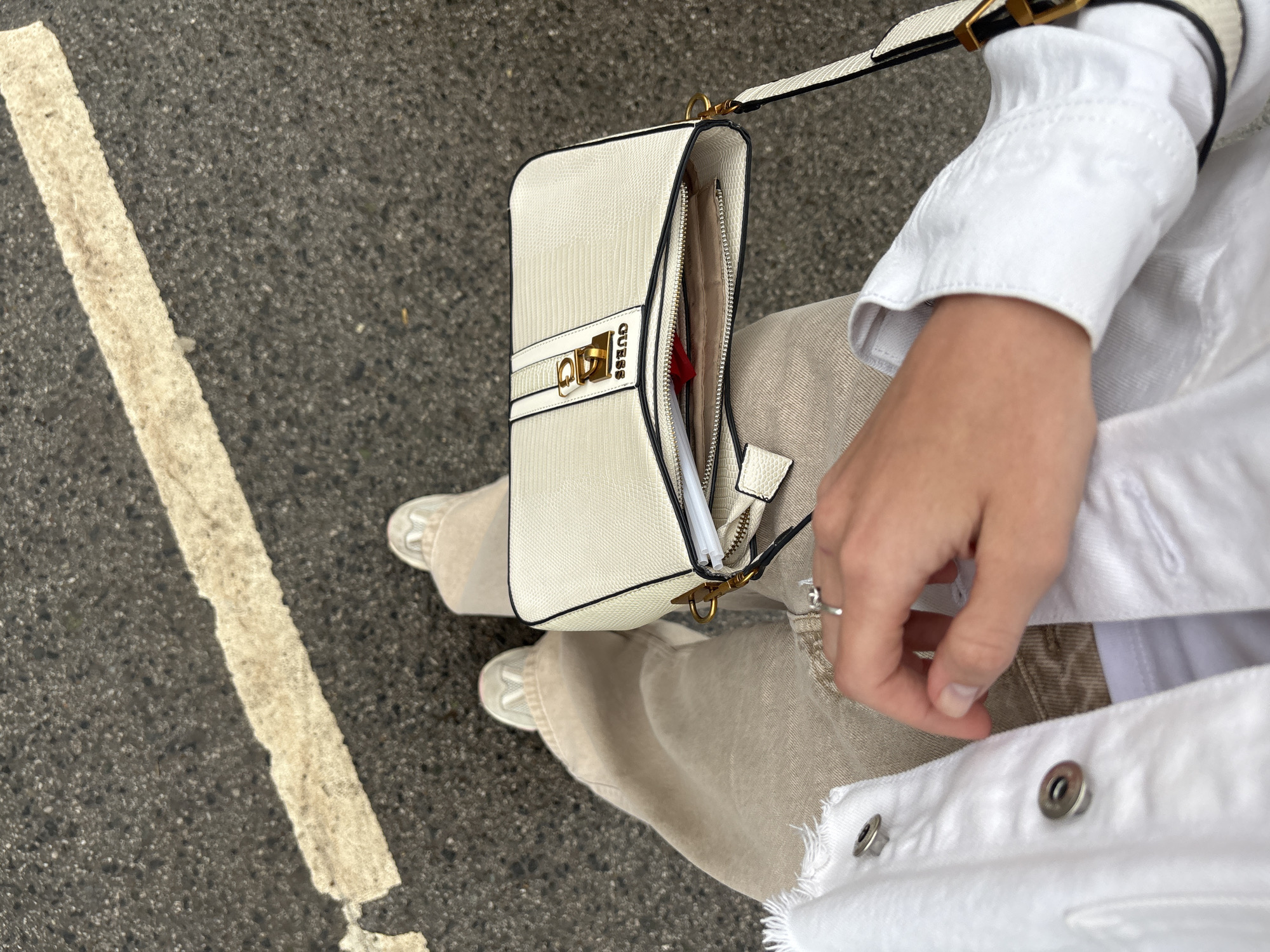 The image depicts a downward, first-person perspective of an individual's lower body and accessories in an outdoor setting. The ground surface is dark gray asphalt with a visible coarse texture, traversed by a worn and irregular off-white painted line. This scene is located in the city of Perm, Russia.
The individual is wearing loose-fitting, wide-leg trousers of a light beige or khaki color. Both feet are shod in light-colored (off-white or light gray) sneakers with white soles, partially visible beneath the trouser hems.
In the lower center-left, the wearer's right hand is visible, resting on their leg. A silver-colored ring with a single clear stone is on the ring finger. A white denim jacket or similar outerwear, with a raw, frayed edge and two silver-colored metal snap buttons, is visible on the left side of the frame. A white undershirt or interior garment is also partially visible beneath the jacket's opening. On the far right, a portion of a white sleeve with a textured pattern and a visible cuff is present.
A rectangular off-white shoulder bag with a textured, scale-like pattern and black piping is positioned centrally on the individual's lap or leg. The bag features a prominent gold-colored metal clasp with the word "GUESS" debossed on it. Its strap is white with black edges, secured by a gold-colored buckle. The bag's main compartment is partially open, revealing a zippered closure, a light beige interior lining, a red object (possibly a wallet), and a white cylindrical item (e.g., pen, lip gloss) inside.