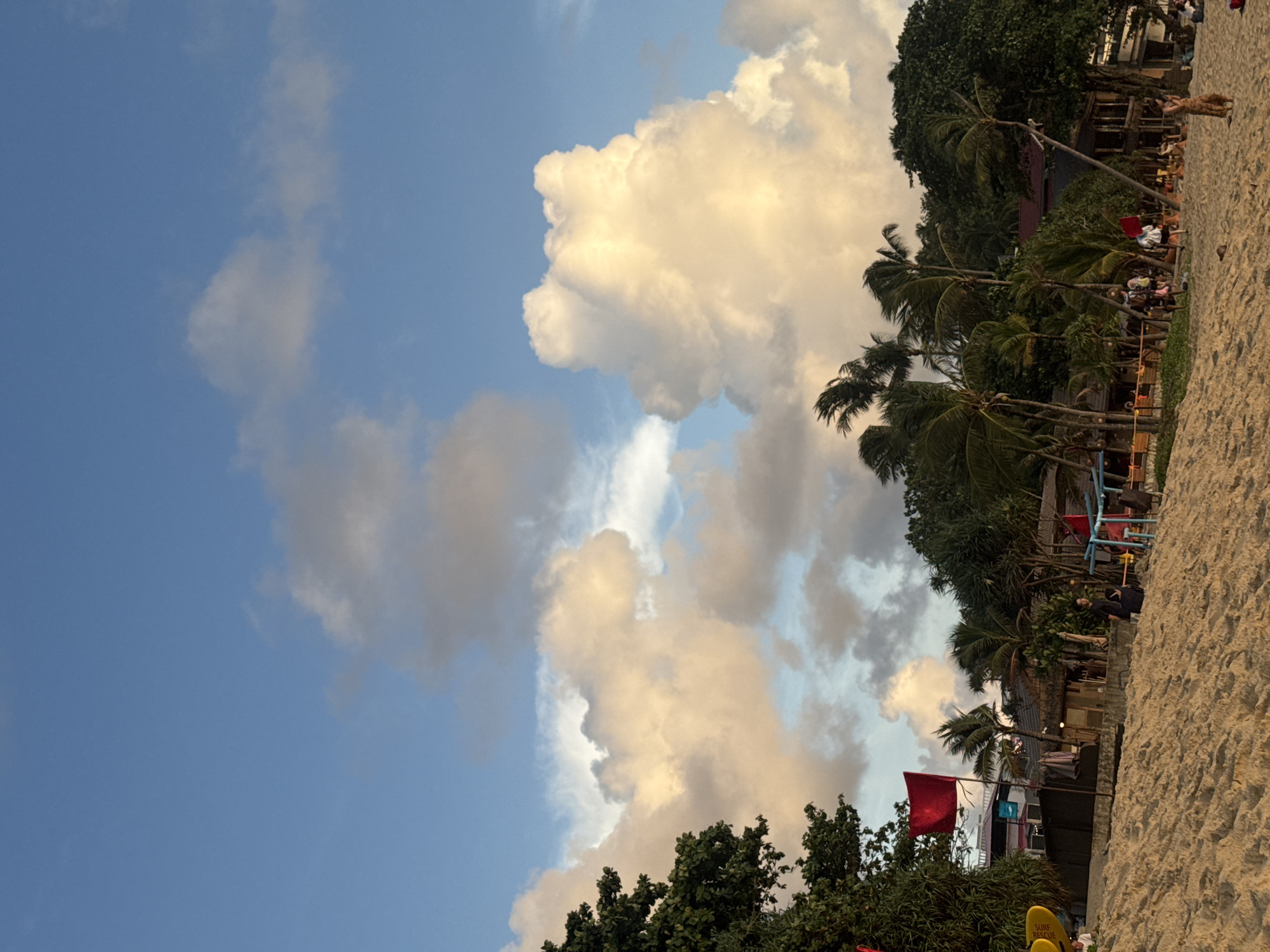 A wide shot captures a beach scene in Karon, Thailand, with a sandy foreground leading to a line of palm trees and tropical foliage. In the background, several low-rise buildings with thatched roofs and wooden structures are visible. A few people are scattered on the beach, some walking and others sitting. Red flags are planted in the sand, and what appear to be surfboards are leaning against a structure on the left side of the image. The sky is filled with large, fluffy white and yellow clouds, illuminated by soft sunlight, suggesting it is either early morning or late afternoon. The overall atmosphere is relaxed and tropical.