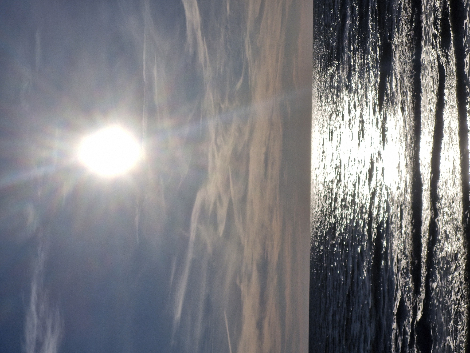 The image shows a wide shot of the Baltic Sea near Karklė, Lithuania, under a partly cloudy sky. The sun is bright and high in the sky, casting a strong glare on the water and creating a shimmering effect on the waves. Wispy clouds, some with airplane contrails, stretch across the sky, tinged with pink and orange from the sunlight. The horizon line is clearly defined, separating the sea from the sky. There are no people or man-made objects visible in the scene.