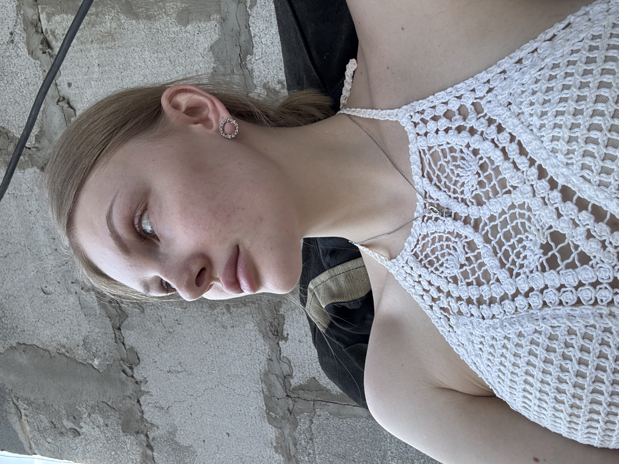 A pale-skinned young woman with fair hair and a silver necklace is positioned against a backdrop of gray concrete. She wears a white crocheted top. The setting appears to be an urban environment. While the image provides no direct evidence of the location being in Perm, Russia, the presence of gray concrete suggests a typical urban environment.