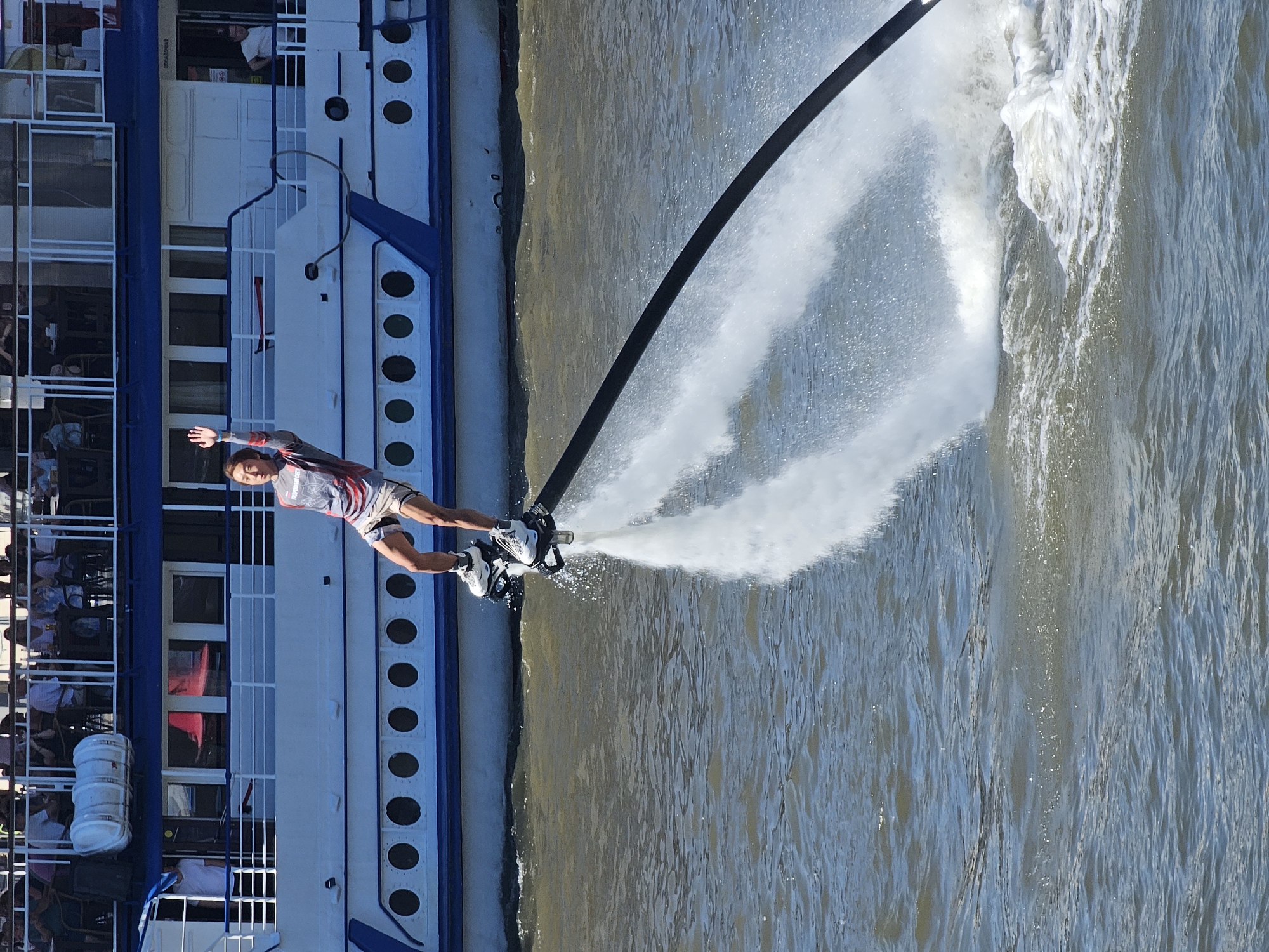 A person is hydroflighting on a flyboard in the Moscow River, in front of a large white boat with blue trim. The person, wearing a grey and red athletic shirt and patterned shorts, is in mid-air, with their right arm raised in a wave. They are wearing white athletic shoes attached to the flyboard, which is directing powerful streams of water downwards, creating a significant spray. The boat appears to be a passenger vessel, with multiple decks and windows, and people are visible on its upper deck. The water is choppy, reflecting the sunlight. The background shows the cityscape of Moscow, Russia.