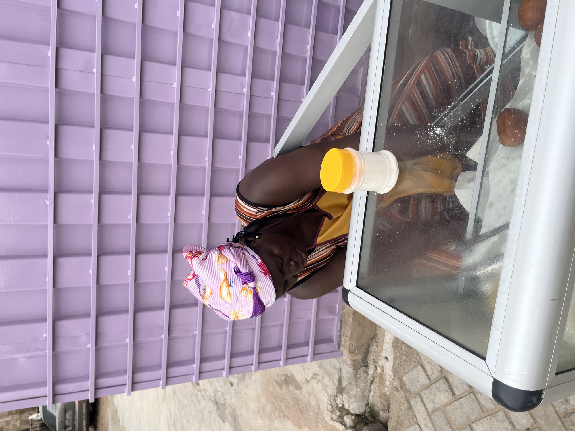 A woman, wearing a pink and floral headscarf and a striped dress over a yellow apron, is looking down into a glass-topped display case. She is positioned to the left of a white plastic container with a yellow lid. The display case contains what appear to be fried dough balls. The background consists of a textured, purple metal wall and a paved walkway. The scene appears to be outdoors in an urban environment, likely in Accra, Ghana, given the context.