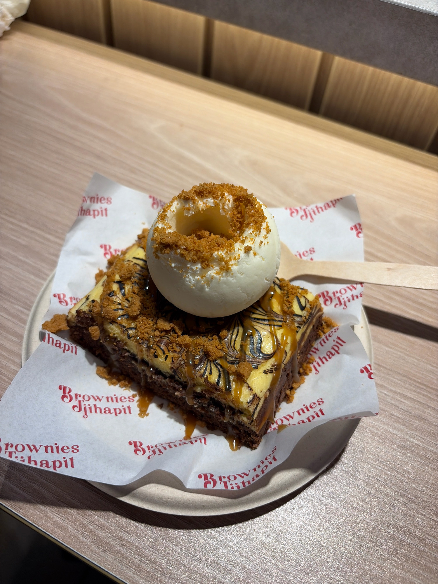 A close-up, overhead shot shows a single slice of cheesecake brownie served on a white paper wrapper with red branding that reads "Brownies Jjihadit". The brownie is topped with a scoop of vanilla ice cream, crumbled cookie pieces, and a caramel drizzle. The dessert is placed on a light-colored plate, which rests on a wooden table. A wooden spoon is visible on the right side of the plate. The overall lighting is bright and suggests an indoor setting, possibly a cafe. The branding on the wrapper, "Brownies Jjihadit," indicates the potential location is Bandung City, Indonesia, as the establishment with this name is located there. There are no people visible in the image.