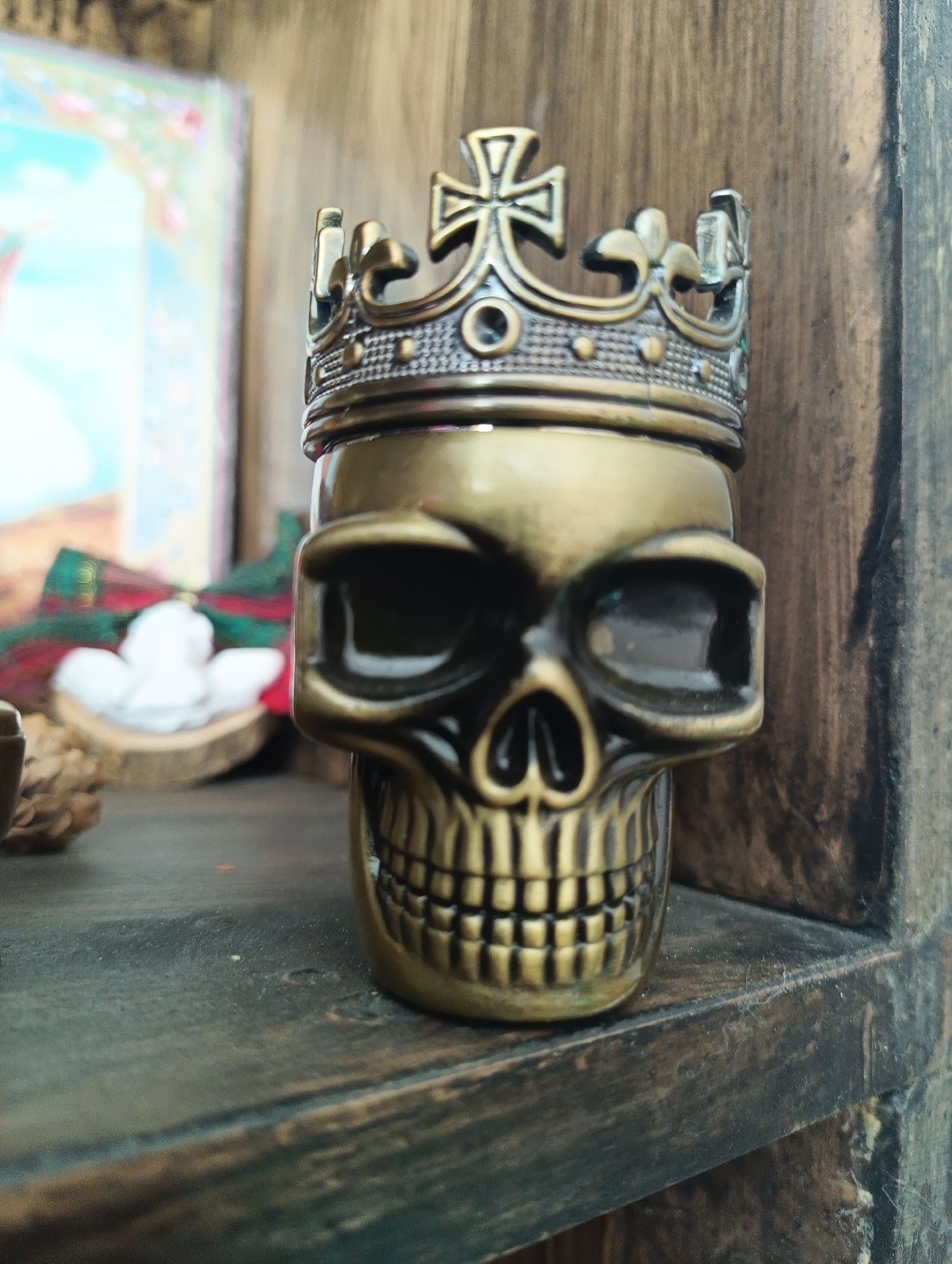 A close-up, eye-level, outdoor, daylight shot shows a metallic skull with a crown on its head. The skull is gold in color, with black accents in the eye sockets and nose area. It has a detailed mouth with visible teeth. The crown is ornate and metallic, featuring a cross at the top and intricate detailing. The skull is positioned on a dark wooden shelf, which appears to be part of a rustic or aged structure. In the background, a blurred image of what looks like a framed artwork is visible, featuring soft colors like pink and blue. To the left of the skull, a red and green plaid ribbon is tied in a bow, behind which a small, white, angel-like figure is visible. The overall environment suggests a decorative display, possibly within a home or shop in Karnobat, Bulgaria.