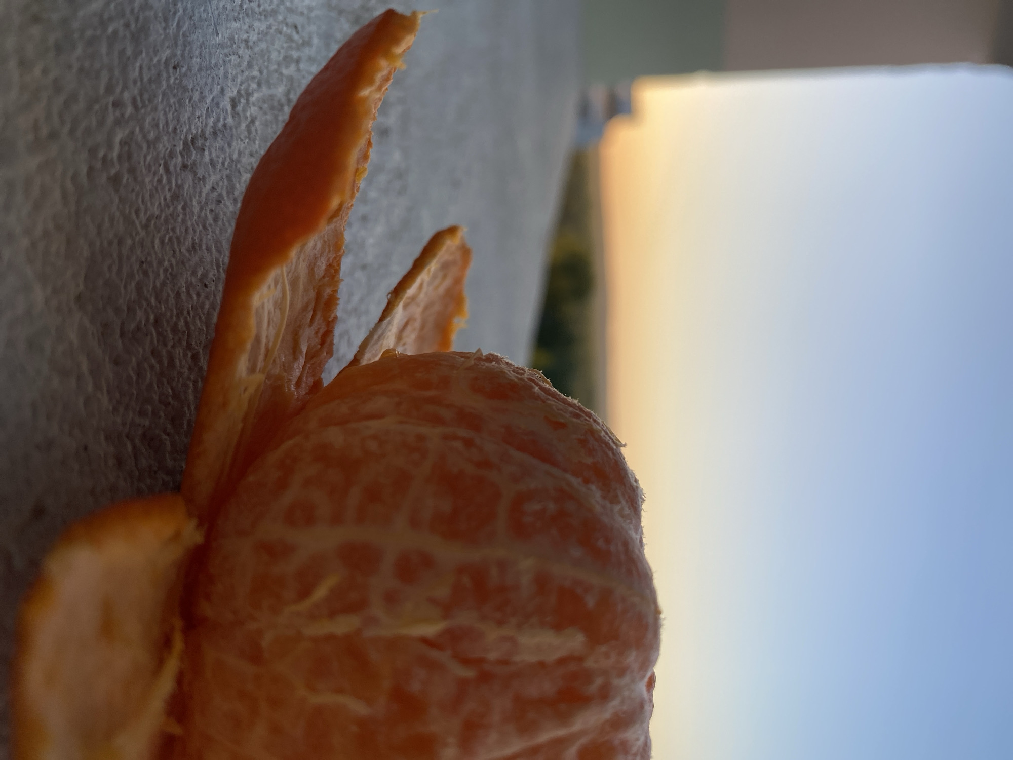 A close-up, slightly out-of-focus, indoor shot of a peeled tangerine. The tangerine sits on a textured, light-colored surface. Portions of the tangerine's peel are partially detached, revealing the fruit beneath. In the background, blurred colors suggest an outdoor view of a building against a light sky, possibly near sunset. The location is unspecified.