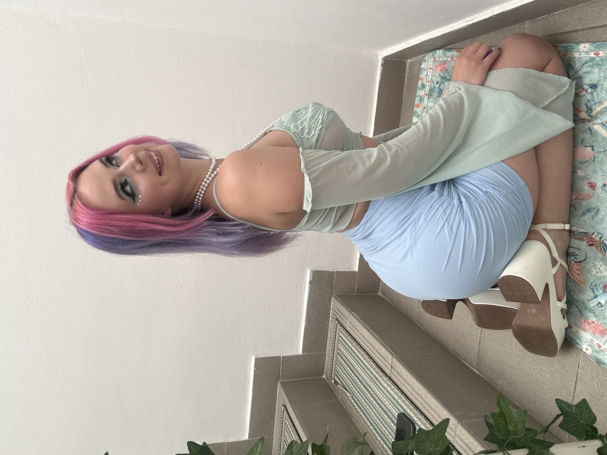A woman with pink and purple hair, wearing a light green top, a blue skirt, and white high-heeled sandals, is sitting on the steps. The steps are carpeted with a patterned rug and bordered by plants. The location is in the city of Zagreb, Croatia.