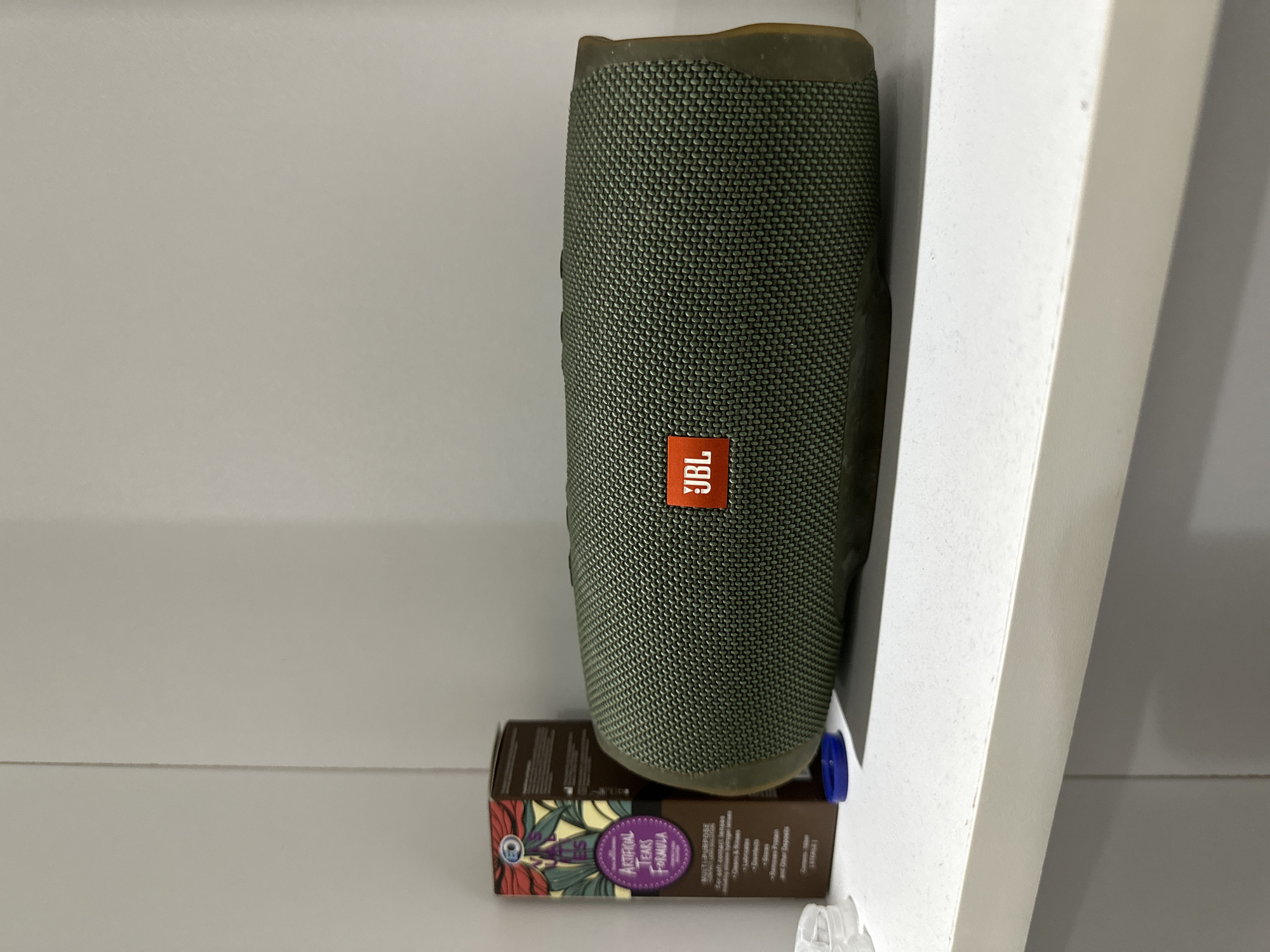 A green JBL speaker is standing vertically on a shelf. The speaker is sitting on top of a box labeled "ARTIFICIAL TEARS FORMULA". The shelf is white and appears to be part of a larger shelving unit. The background is a plain white wall. No people are visible. It is not possible to determine if this image was taken in Davao City, Philippines, or anywhere else.