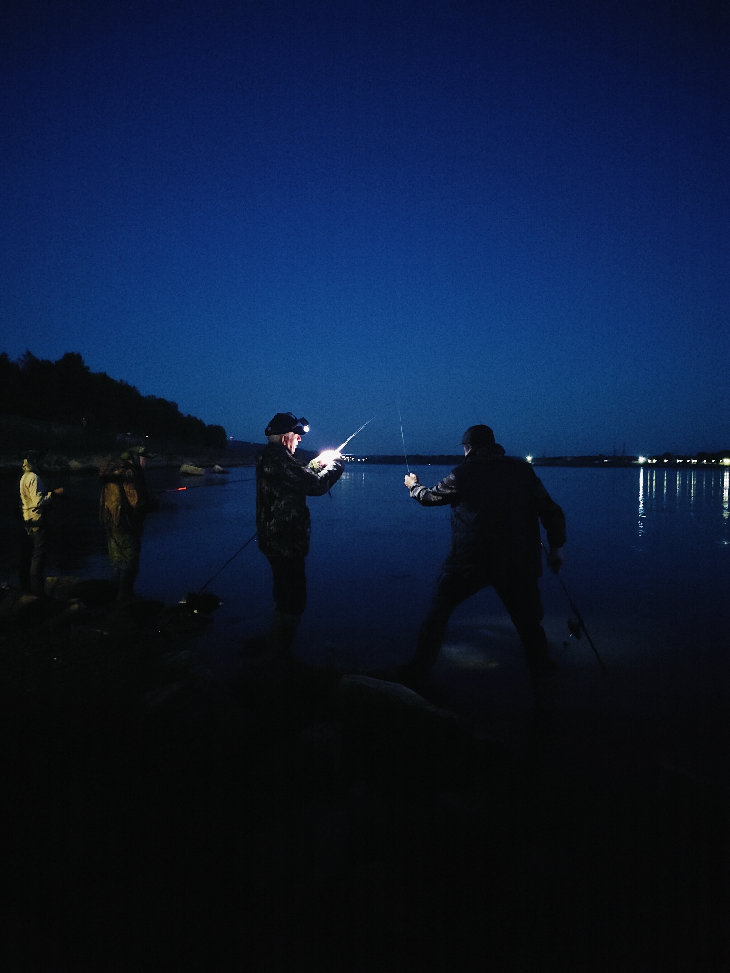 Four individuals are fishing at dusk on the bank of the Kama River in Chaikovsky, Russia. Two men in the foreground are actively fishing, with one wearing a headlamp that illuminates his fishing rod. The water is calm, reflecting the dim lights of a distant town. The sky is a deep blue, indicating either early morning or late evening. The bank is rocky and covered in sparse vegetation.