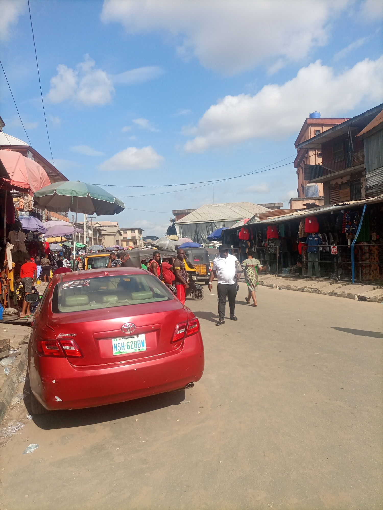 The image depicts a street scene in the Fegge area of Nigeria. A red Toyota Camry is parked on the right side of the road, facing away from the camera. The car's license plate reads "NSH-628BW". The street is lined with market stalls on both sides, displaying various goods, primarily clothing. Several people are walking and interacting within the market. Some individuals are dressed in casual attire, while others appear to be vendors or customers. In the background, there are buildings, some of which are multi-story structures with balconies. The sky is blue with scattered white clouds. The overall atmosphere suggests a busy, active marketplace environment.