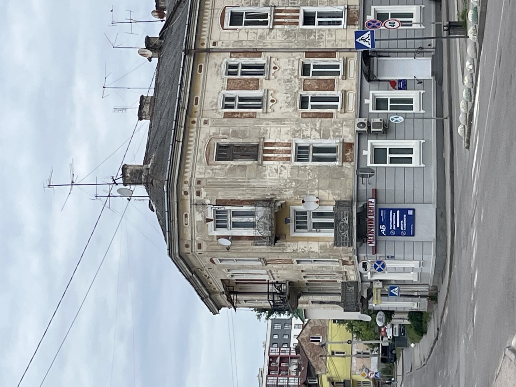 The image shows a street view in the city of Drohobych, Ukraine, focusing on the corner of an old, weathered apartment building with ornate architectural details. The building is a pale yellow with beige trim, showing signs of age and peeling paint. It has multiple windows with white frames, some with balconies. Above the building, several television antennas and satellite dishes are visible against a cloudy sky. To the left of the main building, there are glimpses of other, more modern buildings, including a taller structure with red and gray elements. A yellow building is also visible further down the street.
At the street level of the corner building, there is a gray facade with a sign in Cyrillic that reads "МЕДИЧНИЙ ЦЕНТР" (Medical Center). Several blue and white street signs are posted, including a pedestrian crossing sign, a circular sign with a diagonal cross, and a circular sign with the number "40" indicating a speed limit. A white door serves as an entrance to the medical center. The street in the foreground is paved, with visible crosswalk markings and concrete bollards along the curb. A dark-colored van or SUV is parked on the street to the left. The overall atmosphere is overcast and somewhat subdued.