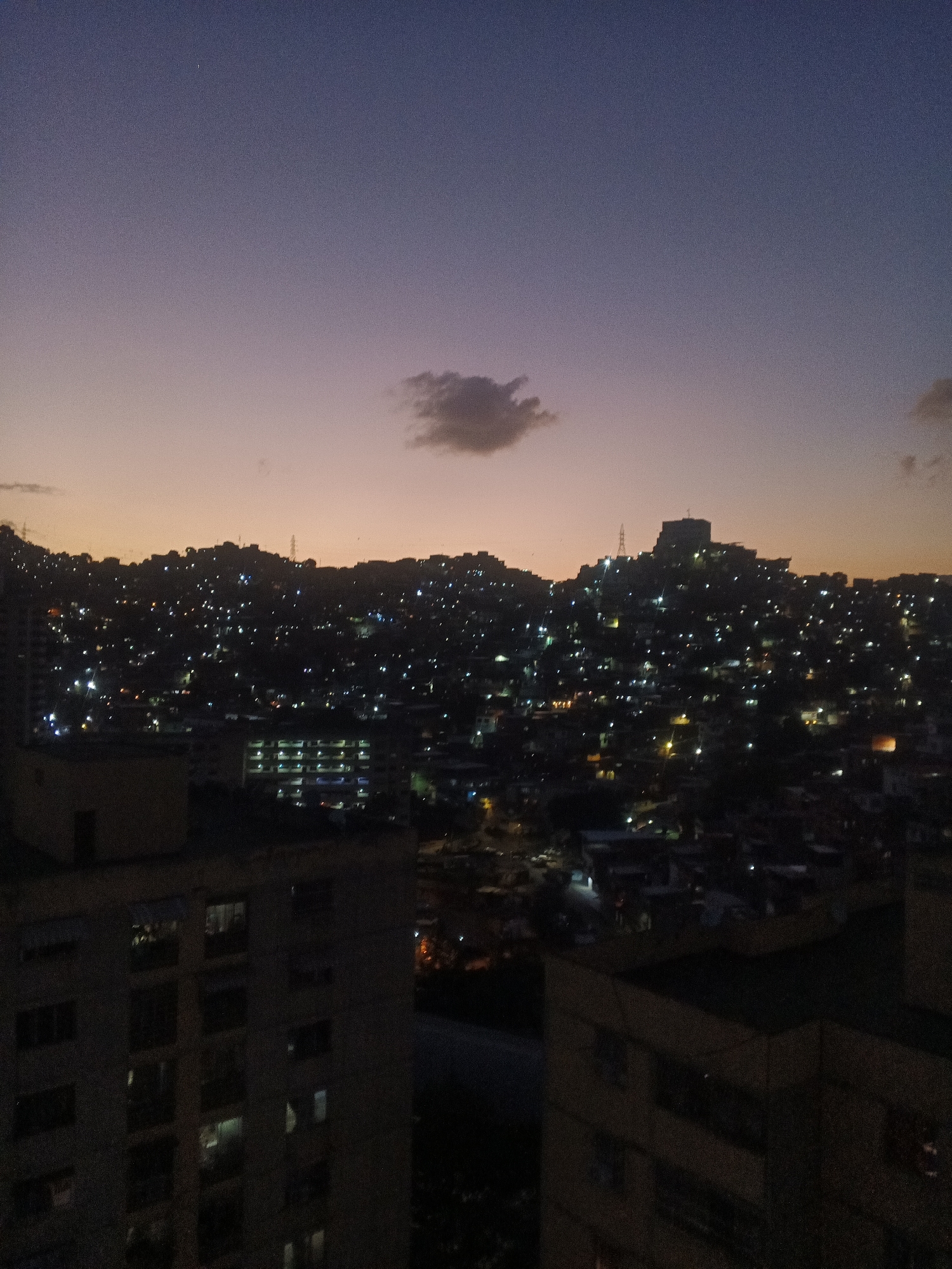 The image depicts an evening cityscape, likely Caracas, Venezuela, given the presence of densely packed housing on the hills. The foreground features the rooftops of several multistory buildings. In the midground, a hillside is covered in numerous closely packed houses, illuminated by scattered lights. A solitary cloud floats in the sky above the hillside. The sky exhibits a gradient of colors, transitioning from a dark blue at the top to a light orange near the horizon. The scene is dimly lit overall, characteristic of twilight or early evening. There are no visible people or interactions.