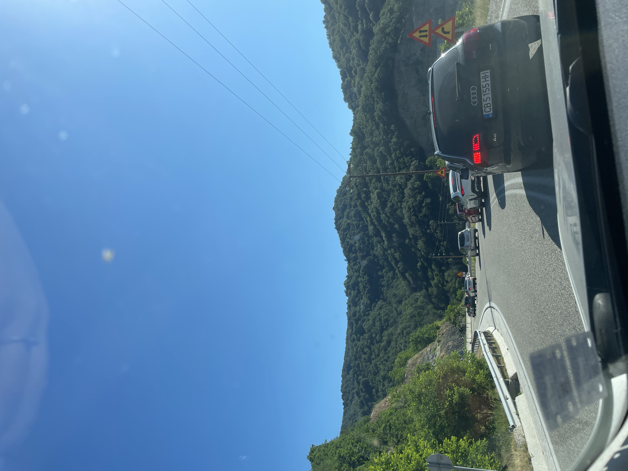 The image shows a line of cars on a road with green hills on either side under a clear blue sky. The camera is inside a vehicle, looking forward. The car in the foreground is a black Audi A6 with license plate CB 5155 HH. Traffic signs indicating caution are visible to the right of the road. Power lines stretch across the sky. The overall scene suggests a journey through a rural or mountainous area. The location is identified as Unknown, Greece.