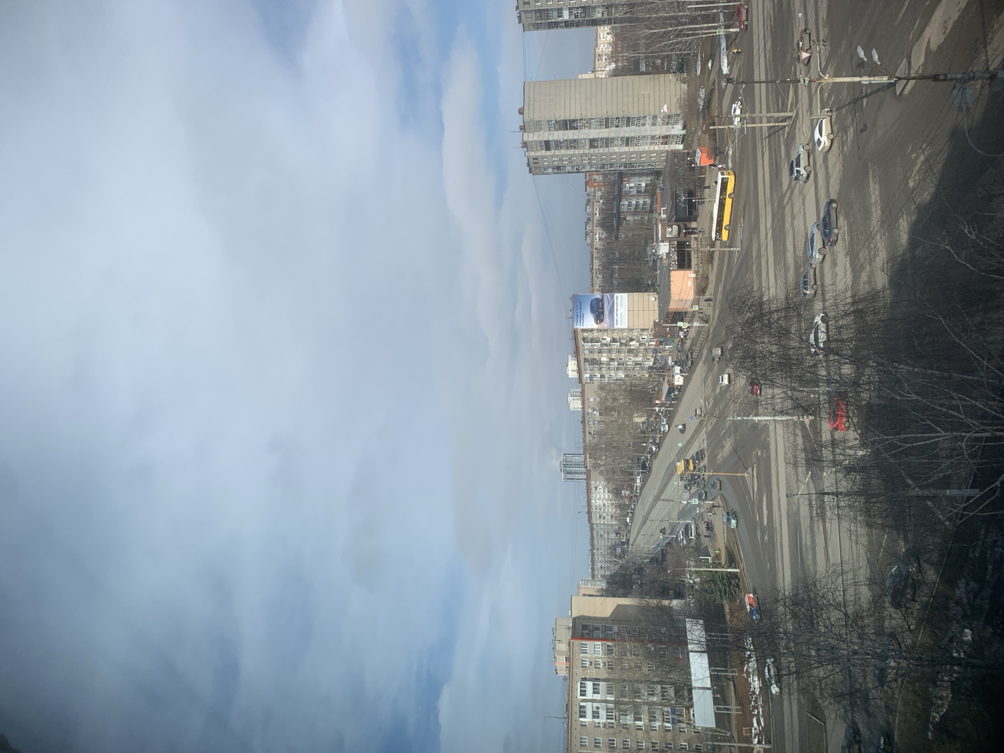 The image depicts a cityscape in Izhevsk, Russia, viewed from an elevated position. The right side of the image shows a busy road with multiple lanes of traffic, including cars and a yellow bus. Buildings of various heights line the streets, with a large billboard visible on one of them. Bare trees are interspersed along the roadside. The left side of the image is dominated by a sky filled with white and blue clouds.
