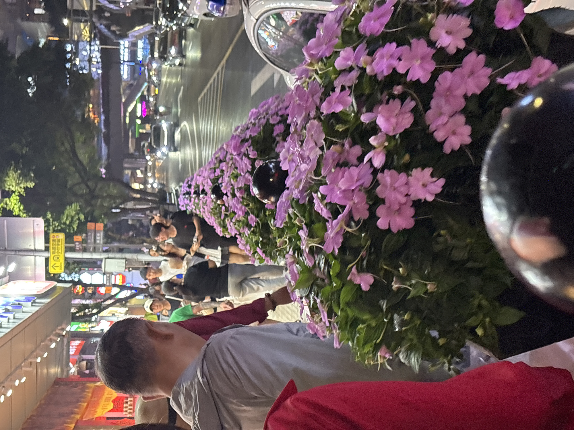 The image shows a street scene at night in Yuzhong District, China. The road is lined with planters containing purple flowers. Several people are walking on the street. A man wearing a gray shirt and red undershirt is in the foreground. Storefronts with neon signs and building lights illuminate the background.