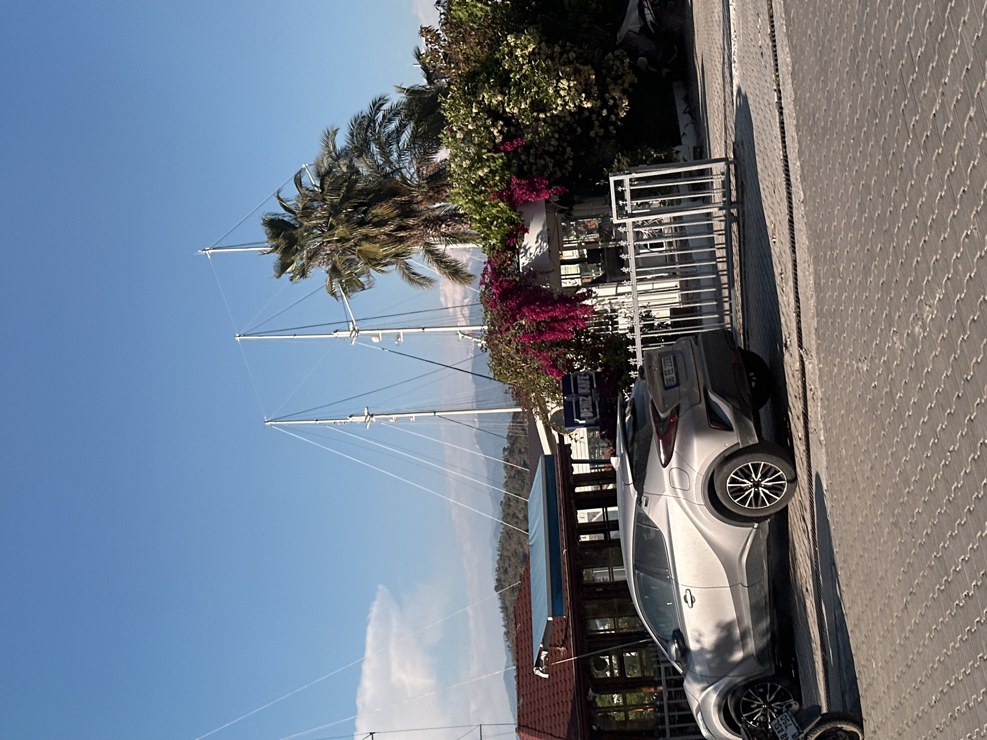 The image captures a sunny outdoor scene in Fethiye, Turkey, depicting a street or promenade alongside a marina. In the foreground, a silver car, possibly a compact SUV, is parked on a light grey, patterned paver sidewalk. The car's front license plate displays "06 EE 2339". Long shadows are cast on the pavement, suggesting either late morning or early afternoon.
To the left of the parked car, the tall white masts and rigging of several sailboats are prominently visible, indicating a harbor or marina just beyond the promenade. Further back, across a body of water, distant mountains rise under a clear, bright blue sky with a few wispy clouds.
Lining the edge of the promenade are lush green trees, including several tall palm trees. Vibrant pink bougainvillea and other flowering bushes add splashes of color. Partially visible behind the foliage are buildings with dark windows and a brown tiled roof, suggesting a cafe or restaurant. A blue sign with white lettering, reading "DENİZ KAFE" (Turkish for "Sea Cafe"), is affixed to one of these structures. White metal railings are also present, bordering the properties. No people are visible in the frame.