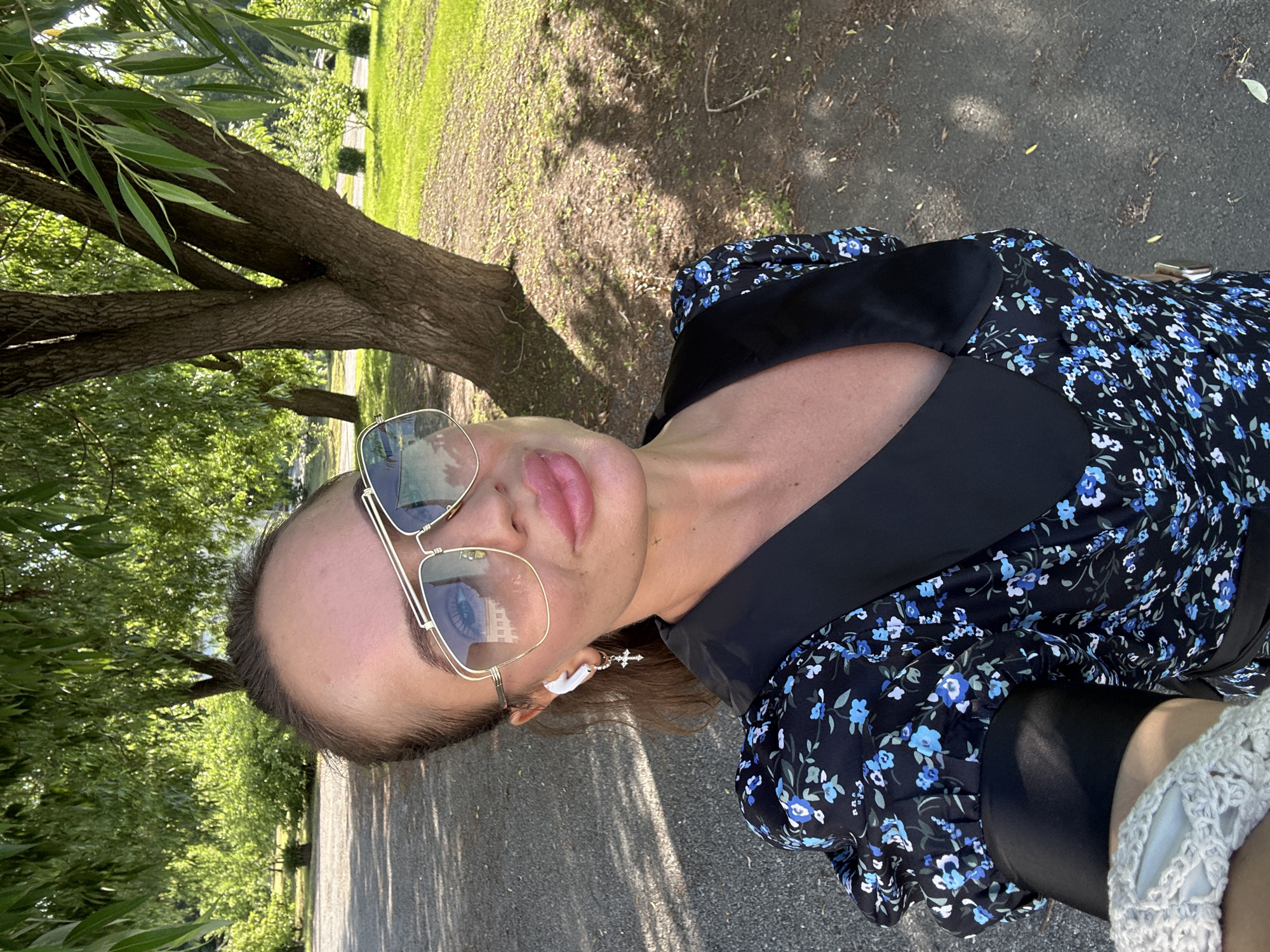 The image shows a woman taking a selfie outdoors. She is wearing a black dress with a blue floral pattern, large gold-rimmed sunglasses, and white wireless earbuds with a cross-shaped charm on one ear. The background consists of a paved path, trees, and a grassy area, suggesting a park or outdoor recreational space. The lighting indicates it is daytime. The image does not provide enough visual information to determine the specific location within the city of Duvankul', Russia.