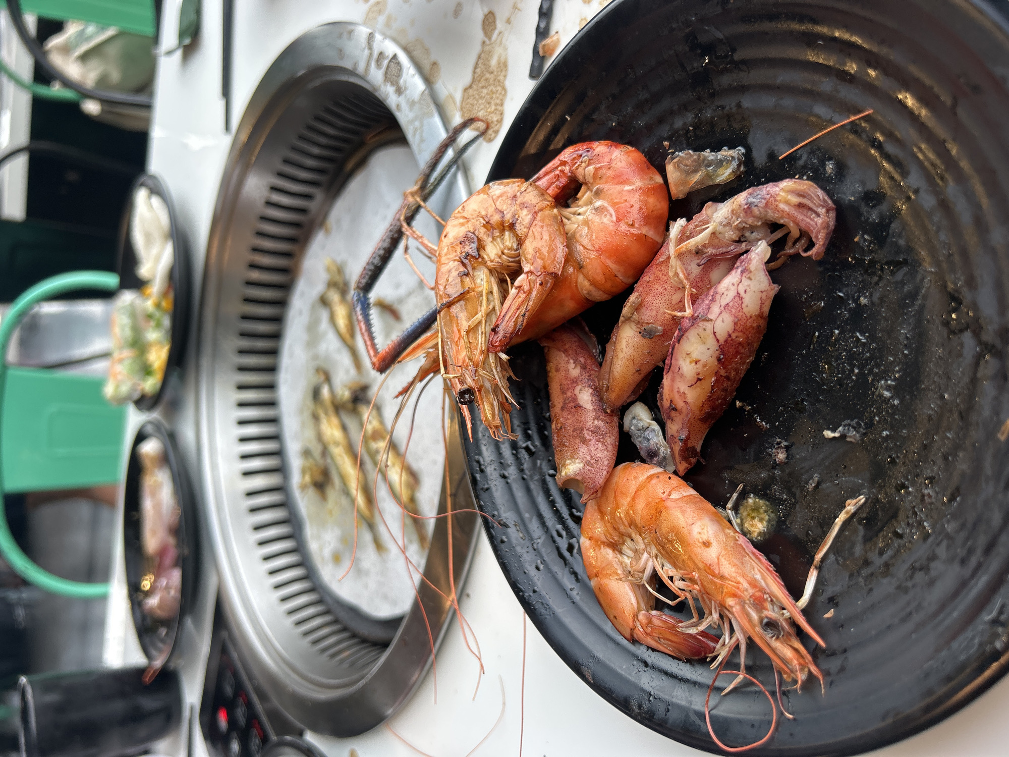 An overhead shot captures a selection of grilled seafood on a table in Nha Trang, Vietnam. In the foreground, a round black plate holds several cooked or partially cooked large prawns, distinguished by their orange-red shells and some visible charring. Also on the plate are pieces of grilled squid, showing a similar cooked texture and coloration. The plate appears used, with some liquid and residue from the seafood.
In the background, to the left, is a circular metal grill with grates, indicating a cooking surface. On this grill, several smaller, elongated fish, possibly dried anchovies or similar, are seen grilling. The grill unit itself is integrated into a white surface, likely a table or countertop. A glimpse of another cooking station or surface with a green-handled utensil or detail is visible further to the left. No people are visible in the frame, and there is no discernible text. The image focuses entirely on the food and cooking setup, suggesting a meal or food preparation activity.