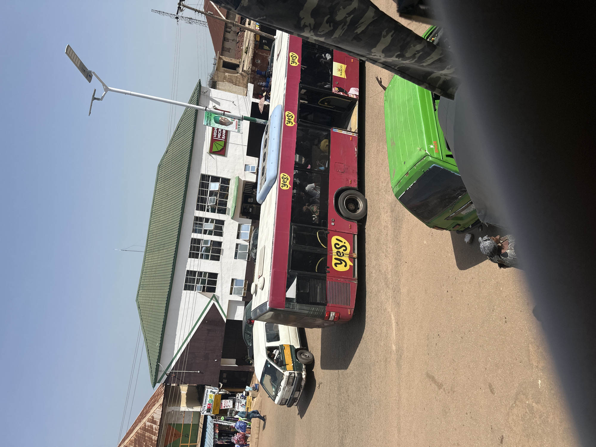 A street scene in Bukuru, Nigeria shows a maroon and red bus with "YES!" written on its side in yellow lettering. The bus has tinted windows and appears to be stopped. To the left of the bus is a white and green van, also stopped, with a person visible inside. In the background, there is a building with a green roof and white walls, and another building with a rusty metal roof. Power lines are visible across the sky. A green vehicle is partially visible in the foreground. The ground is paved.