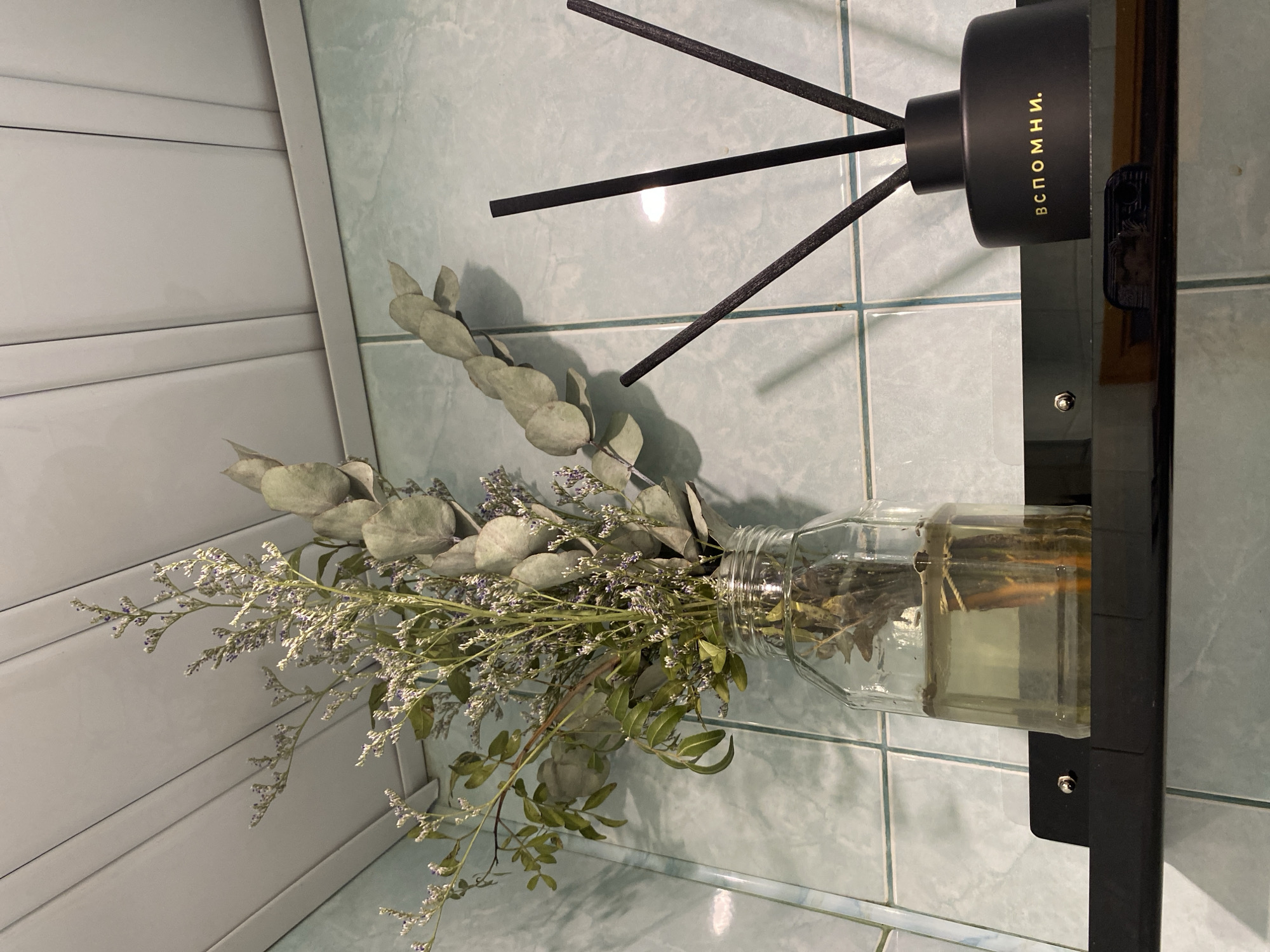 A still-life photograph depicts a clear glass vase with dried flowers and eucalyptus stems, alongside a black diffuser with black reeds, placed on a black shelf attached to a blue-tiled wall. The diffuser bottle is labeled "Вспомни." The location is implied to be Krasnoyarsk, Russia based on the Cyrillic text.