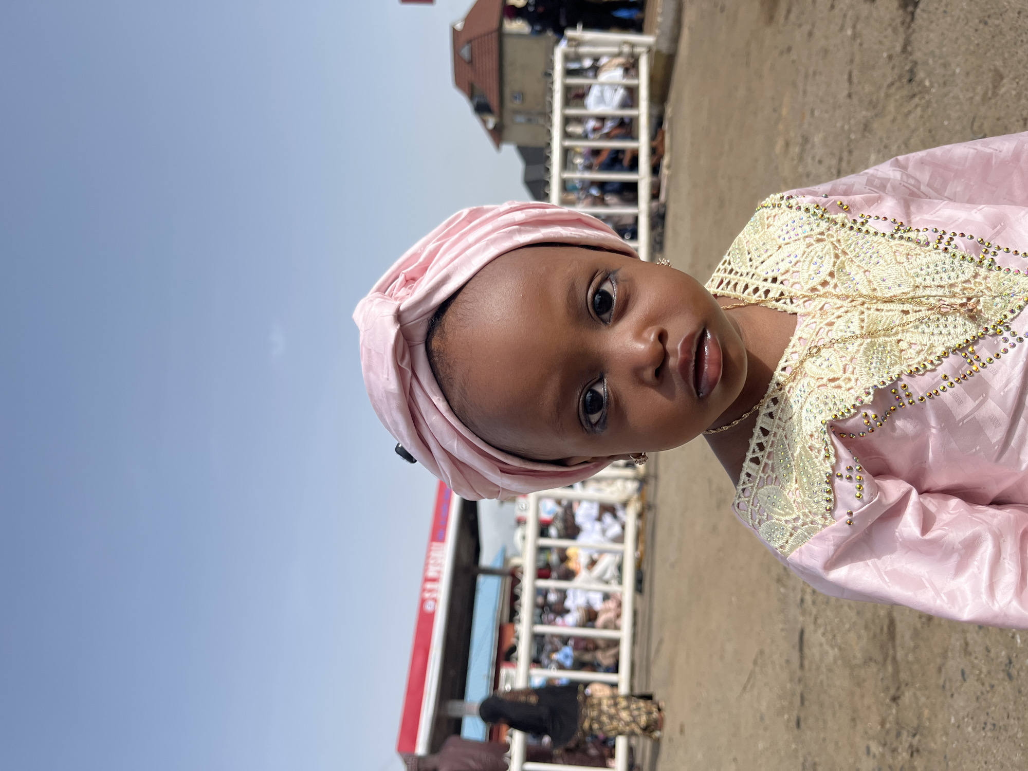 A young child, with brown skin, is the central subject of the photo. The child is wearing a light pink head covering and a pink dress with ornate golden trim. The child has dark eyes and a focused gaze.
The background reveals a crowded area, possibly a public event, near what appears to be a gas station, recognizable by its sign. There's a railing separating the child from the crowd. Other people are visible in the distance, some wearing traditional garments. The environment suggests a sunny day in an urban setting. The city of Jos, Nigeria, can not be accurately determined from the image.