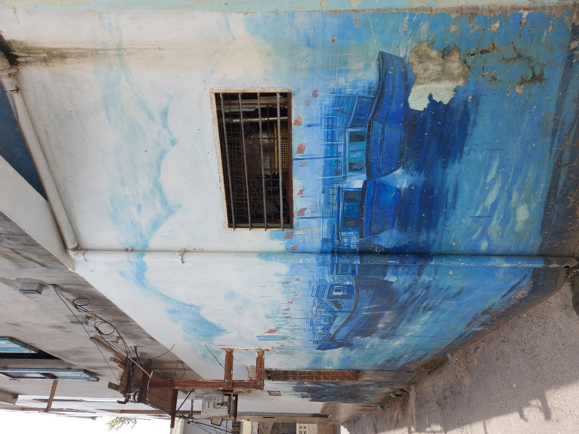 The image depicts a mural of blue fishing boats on a building wall in a narrow alleyway in Da Nang, Vietnam. The mural covers a large portion of the wall, extending from the ground up to a window with a metal grate. The boats are depicted in a calm blue sea, with red flags flying from some of them. In the background, a stylized cityscape is visible. The building itself appears weathered, with peeling paint and some signs of rust. The alleyway is paved with a rough surface and is partially shaded. Electrical conduits and an air conditioning unit are visible on the wall of an adjacent building. The overall impression is one of a community living by the sea, with a touch of artistic expression on its everyday environment.