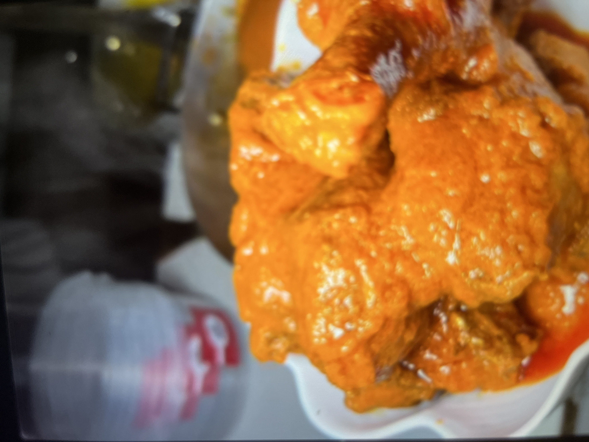 The image shows a close-up of a bowl of buffalo wings coated in a vibrant orange sauce. The wings appear to be tender and glistening, with some flecks of white seasoning visible. The background is blurred, but suggests an indoor setting with containers and possibly other food items. The image does not contain any information that can be used to determine its location in Abuja, Nigeria, or any other specific geographical area. There are no people, actions, or interactions depicted in the image. The focus is entirely on the food.
