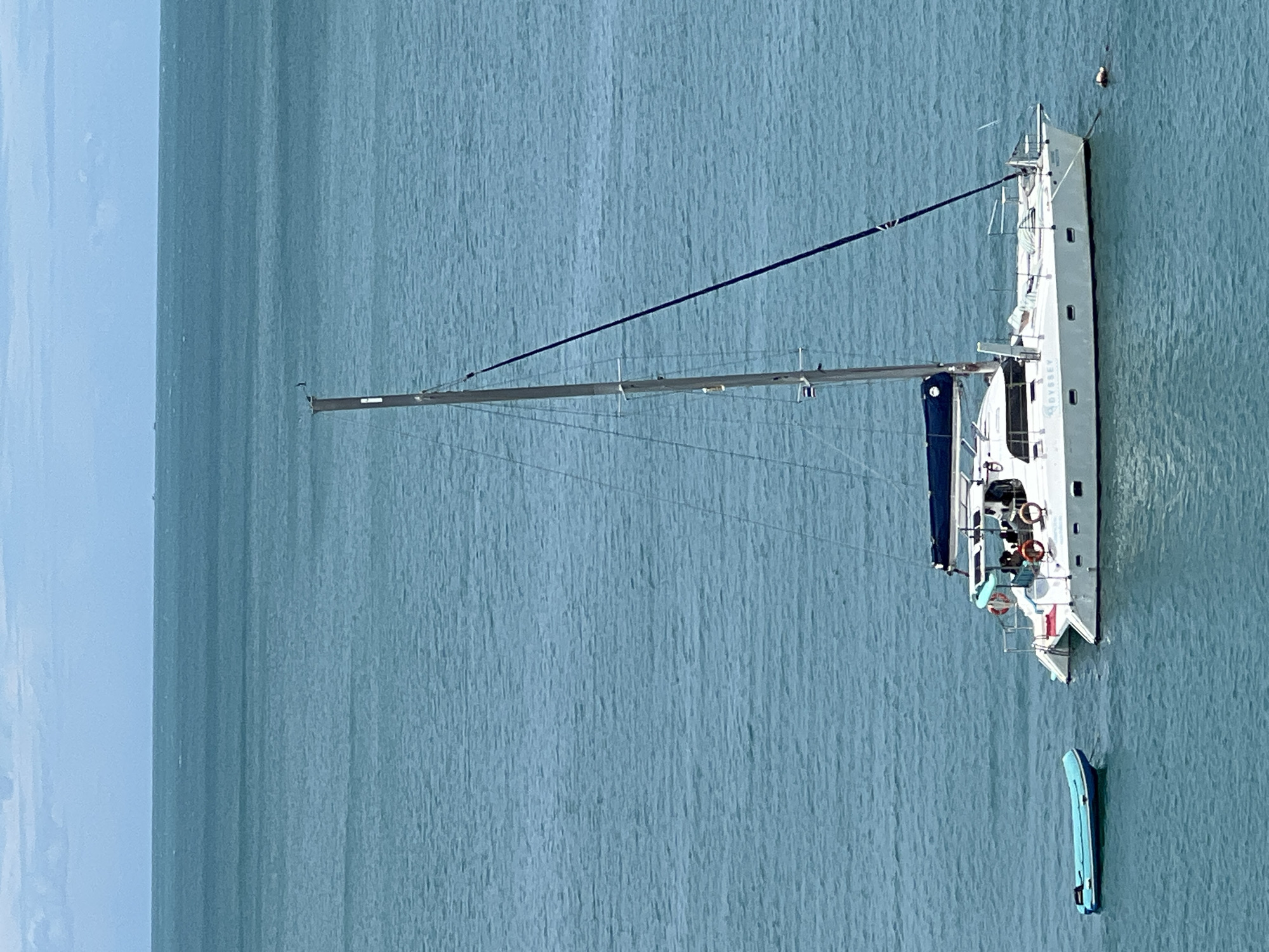 This is an aerial, medium shot of a white catamaran sailing on calm, turquoise water, approximately two miles off the coast of Koh Samui District, Thailand. The catamaran has a single mast with rigging and a dark blue sail cover over the boom. A smaller, inflatable blue dinghy is tethered to the port side of the catamaran. A single person is visible in the water on the starboard side, near the stern, appearing to be swimming or treading water. The ocean extends to the horizon, with faint clouds in the pale blue sky. The name "ODYSSEY" is visible on the hull of the catamaran.