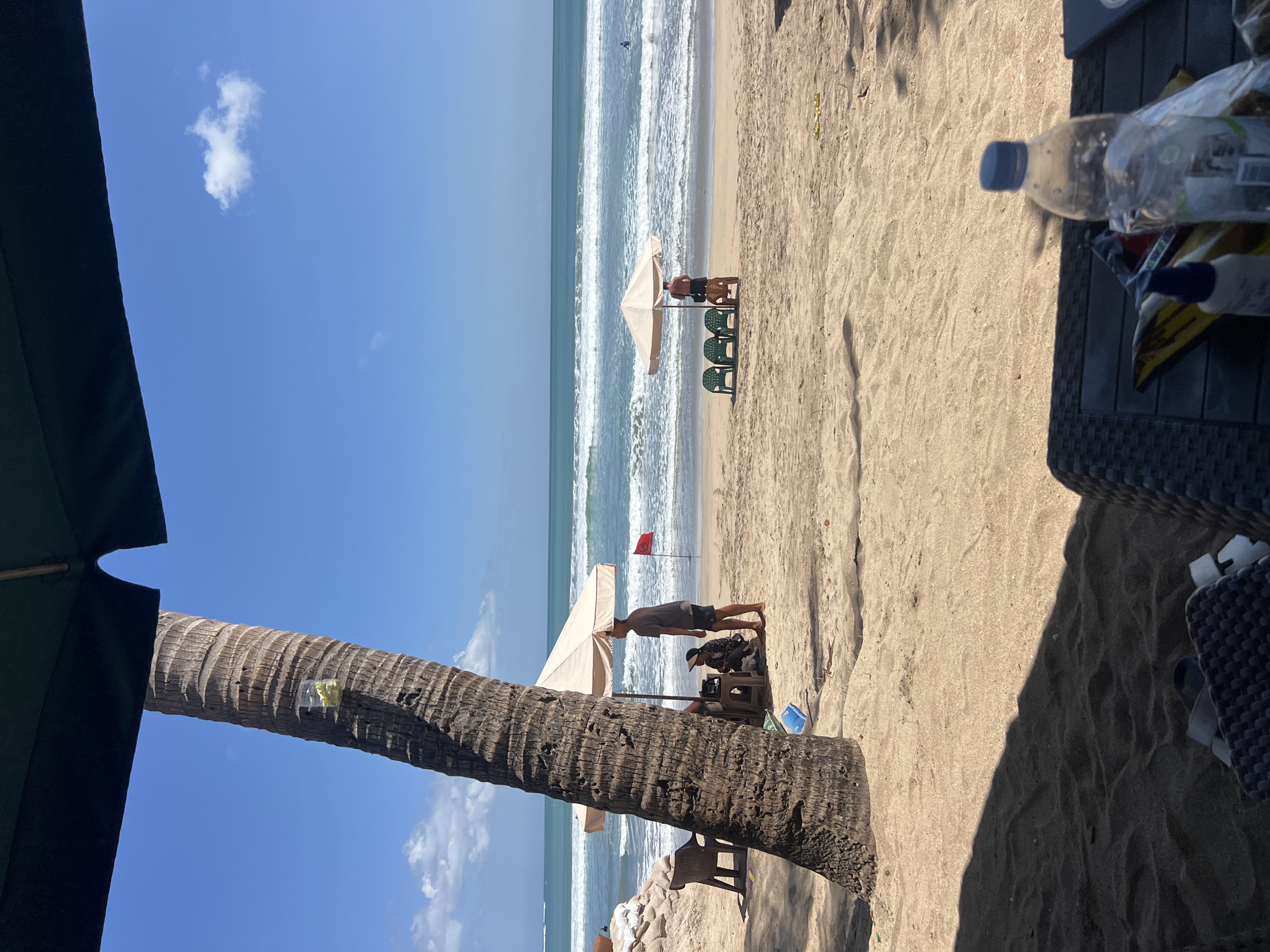 In Kuta, Indonesia, on a sunny day, a beach scene unfolds under a clear blue sky with scattered clouds. The foreground is dominated by the sandy beach, with a large, textured palm tree trunk standing prominently to the left. A dark canopy of an umbrella shades a portion of the sand.
Further back, two beach umbrellas provide shelter for beachgoers. Under the left umbrella, a person sits on a chair next to a table where two other individuals are partially visible. A red flag is planted in the sand nearby. To the right, another umbrella is set up, with four green plastic chairs arranged in front of it. A person stands near the chairs, facing the ocean, while another person is seated further down the beach.
The ocean stretches out to the horizon, with gentle waves rolling onto the shore. A lone figure can be seen swimming in the water. In the distance, a sandy berm or embankment is visible on the left side of the frame. On a table in the foreground, a partially empty plastic water bottle and other items are present. The overall atmosphere is one of a relaxed beach day.