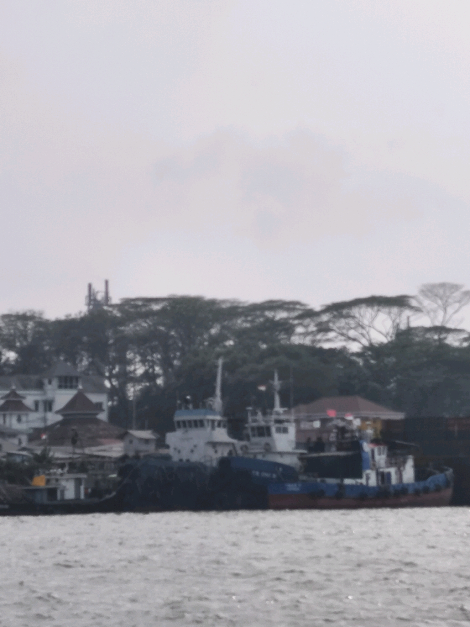 The image depicts a waterfront scene in Palembang, Indonesia, likely along the Musi River. In the foreground, choppy grey water is visible. Dominating the middle ground are several boats. A large, dark hull of a vessel occupies the right side. To its left, a blue and red boat with a white superstructure is moored. Further to the left, a smaller white boat with a yellow detail on its side is also present. Behind the boats, a dense line of trees with lush green foliage forms a backdrop, with some bare branches visible. Several buildings with white walls and red-tiled roofs are nestled amongst the trees. Two tall communication towers are visible above the treeline in the distance. The sky is overcast and grey, suggesting a cloudy day.
