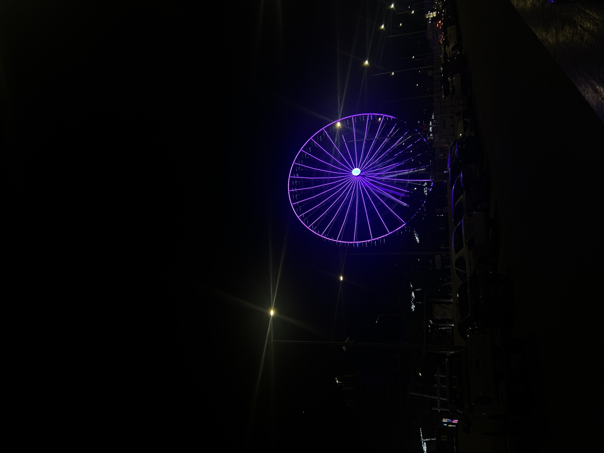 At night in San Bernardino Tlaxcalancingo, Mexico, a Ferris wheel with purple lights stands against the dark sky. The Ferris wheel is lit up, with light reflecting around it. Other structures are on the right, with lights visible. The left side of the image is mostly dark.