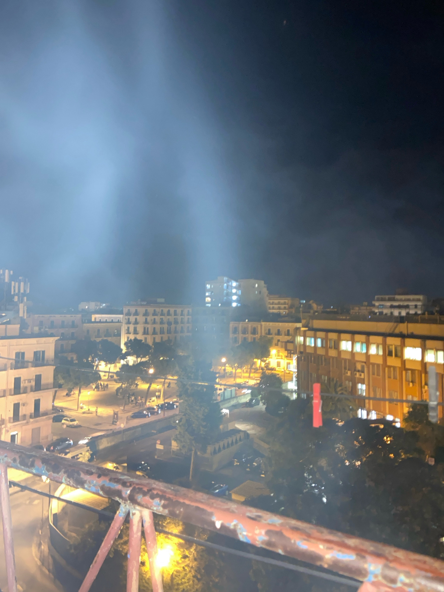 The image shows a nighttime view of an urban area with buildings and streetlights. In the foreground, a rusty metal railing is visible. Beyond the railing, there are multi-story buildings with illuminated windows. Trees and street lights line the streets, where several cars are parked or driving. A hazy, light-colored smoke or fog hangs in the air, obscuring some of the background. The scene appears to be a residential or commercial district in the city of Taranto, Italy, likely taken from an elevated position.