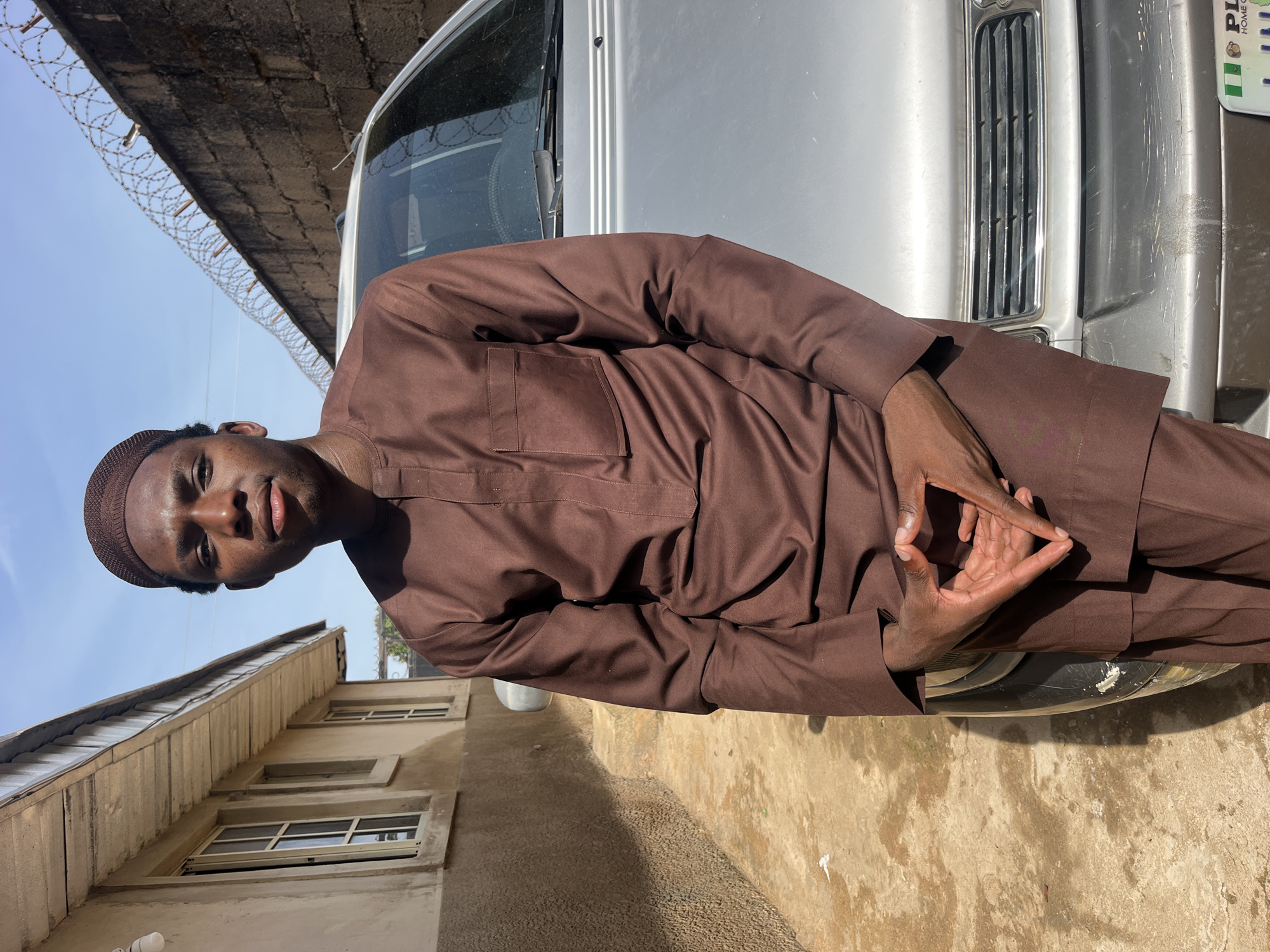 A young Black male stands outdoors, facing the camera, in front of a silver car. He is wearing a brown cap and a matching brown traditional two-piece outfit. His hands are clasped together in front of him, forming a triangular shape. The background shows a building with windows on the left and a brick wall with barbed wire on the right. The ground is paved and appears to be a pathway or courtyard. The lighting suggests it is daytime with bright sunlight casting shadows.