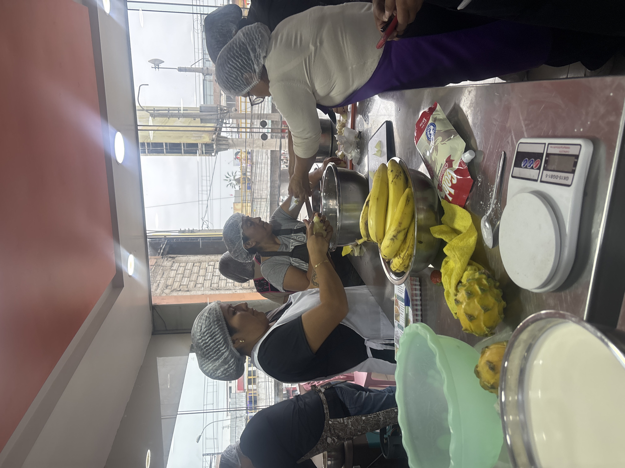 In San Juan de Miraflores, Peru, a group of individuals, predominantly women, are engaged in food preparation in what appears to be a commercial kitchen. They are all wearing hairnets, suggesting a focus on hygiene. In the foreground, a large mint green bowl and a metal bowl filled with a white creamy substance are visible. A digital scale with a white platform sits on a stainless steel countertop, next to a yellow pouch labeled "Colada". Several bananas are arranged in a metal bowl, and two yellow dragon fruits are also present on the counter. The background reveals a brightly lit interior with large windows overlooking an urban outdoor environment. Several people are actively involved in chopping or preparing ingredients, with one woman in the center wearing a white apron and a black t-shirt, holding a knife and working on a food item. Another woman to her right, wearing glasses and a white shirt with a purple apron, is also actively engaged in food preparation. A person on the far left, dressed in black and wearing a patterned apron, is also participating in the activity. The overall scene depicts a collaborative effort in a culinary setting.
