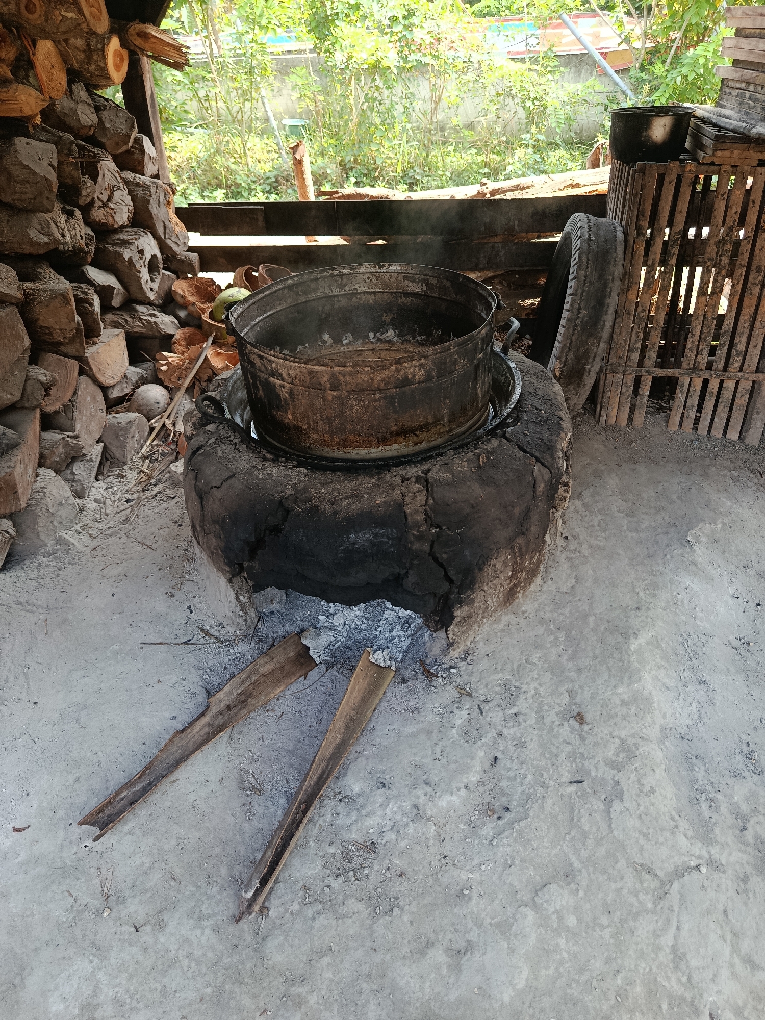 In Sarwadadi, Indonesia, an outdoor cooking scene unfolds. A large, weathered metal pot sits atop a makeshift stove constructed from an old tire and solidified mud, with an opening revealing glowing embers and ash. Two dried palm fronds lie on the dusty ground in front of the stove, likely used as kindling. To the left, a neat stack of chopped firewood adds to the rustic atmosphere. In the background, lush green foliage suggests a natural, possibly rural, setting. The scene is illuminated by natural daylight, casting shadows and highlighting the textures of the materials.