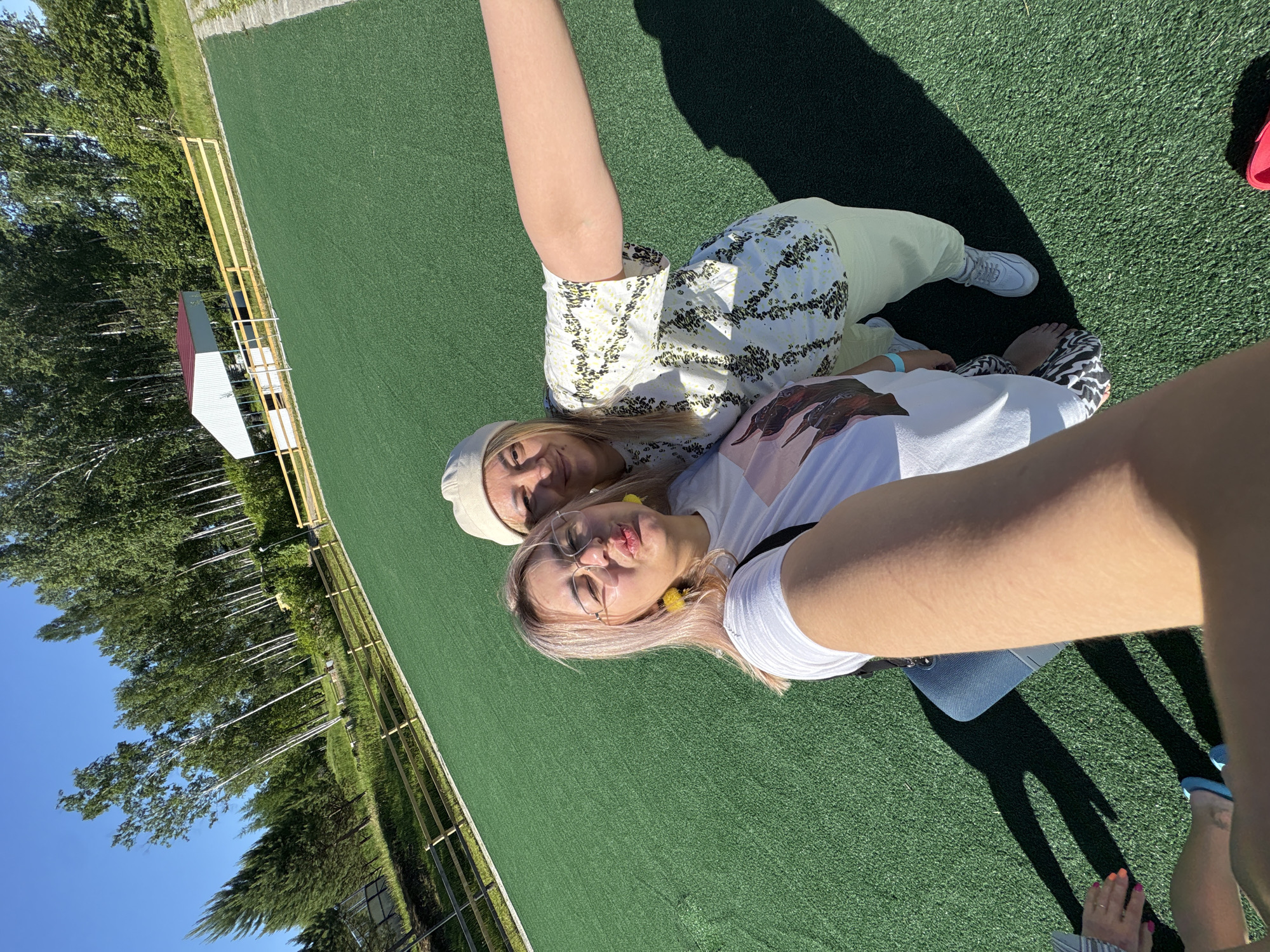 Two young women are taking a selfie outdoors on a sunny day. They are standing on a bright green artificial turf field, possibly a sports field or recreational area. Behind them, there is a light-colored wooden fence and a line of tall, slender trees, characteristic of the boreal forest. In the background, a white structure with a red roof is visible, possibly a pavilion or small building. The sky is a clear, vibrant blue. One woman, in the foreground on the left, is wearing glasses, pink-tinged hair, and a white t-shirt with a graphic print. She is making a puckered-lip expression. The other woman, on the right, is wearing a light-colored beanie and a white t-shirt with a black and yellow abstract pattern. She has her arm extended, holding the camera. The overall setting suggests a pleasant outdoor activity in a natural or semi-natural environment. The specific location in Birobidzhan, Russia, is not visually identifiable from the image content alone.