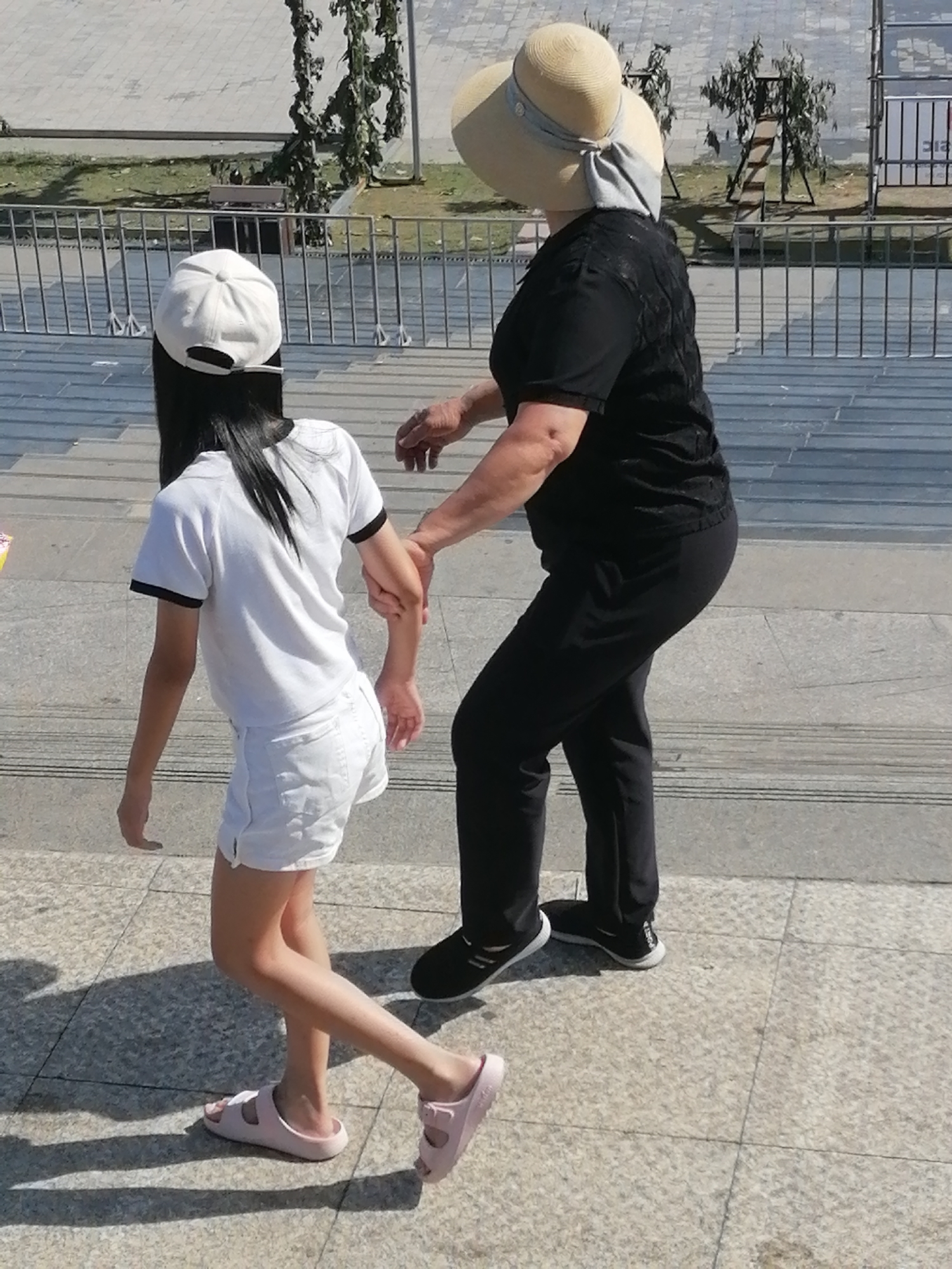 This image depicts two individuals on a paved walkway. On the left, a young person wearing a white baseball cap, white t-shirt, and white shorts is walking. On the right, an older person in a wide-brimmed straw hat and a black shirt is holding the younger person's arm as they walk. The environment appears to be an outdoor urban setting with metal railings and landscaping in the background. The ground is paved with stone tiles. There is no visible indication of the city of Da Lat, Vietnam in the image.