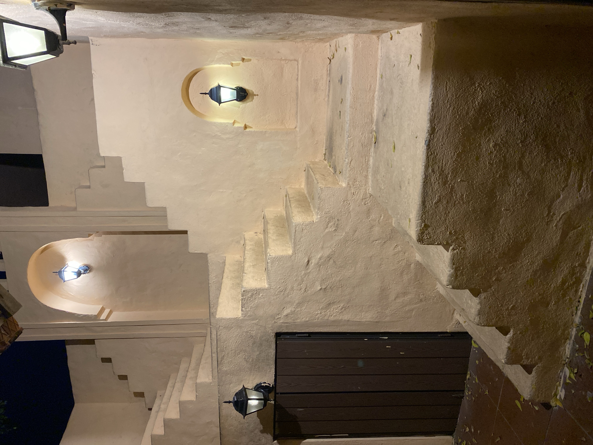 The image depicts a dimly lit outdoor staircase with several lamps illuminating the cream-colored stucco walls and steps. The scene appears to be at night, with the only light sources being the lamps, which cast warm glows. The staircase has a rustic, somewhat weathered appearance, with some fallen leaves scattered on the steps and the tiled floor below. There are at least three visible lamps, one positioned higher up in a recessed alcove, another further down in a similar alcove, and a third one near a dark, wooden-paneled door at the bottom. The architecture suggests an older building, possibly in a courtyard or an alleyway. The overall mood is tranquil and slightly mysterious due to the low lighting and shadows. There is no visible text in the image. The location context is Đà Nẵng, Vietnam.