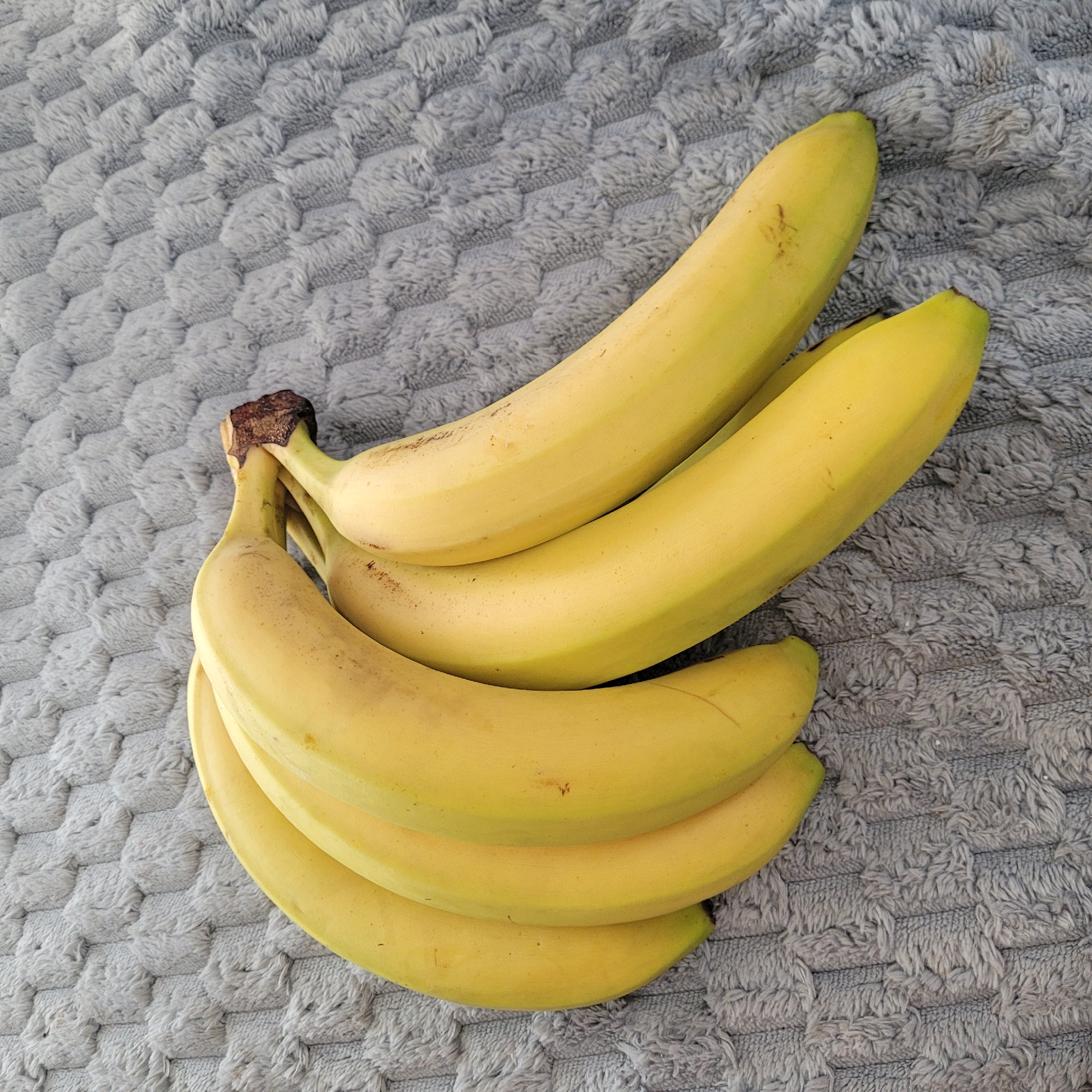 A bunch of six yellow bananas rests on a textured grey fabric surface. The bananas are arranged in a slight curve, with their stems pointing towards the upper right of the frame. The fabric has a raised, hexagonal pattern with a soft, fuzzy texture. There are no people or discernible activity in the image. The scene is indoors, and the lighting appears to be ambient. The image does not provide any information or context related to the city of Jūrmala, Latvia.