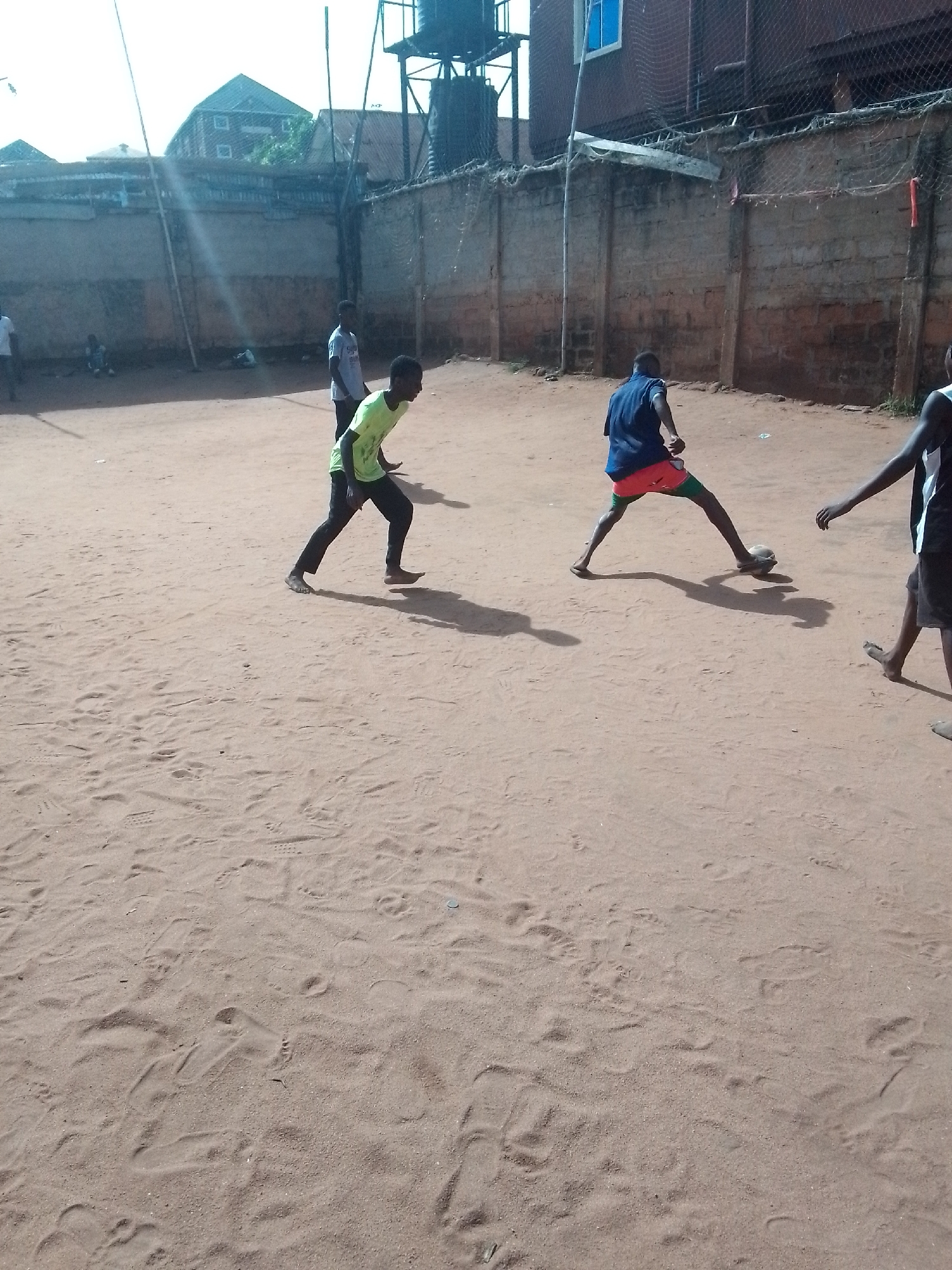 The scene depicts a group of young men playing soccer on a sand-covered field in an outdoor location. The field is surrounded by concrete walls, with a building and water tower visible in the background. There are at least four individuals actively engaged in the game. The player on the right is wearing a blue shirt, orange shorts, and green knee socks, while the player on the left is wearing a light green shirt and black pants. The location is Unknown, Unknown.