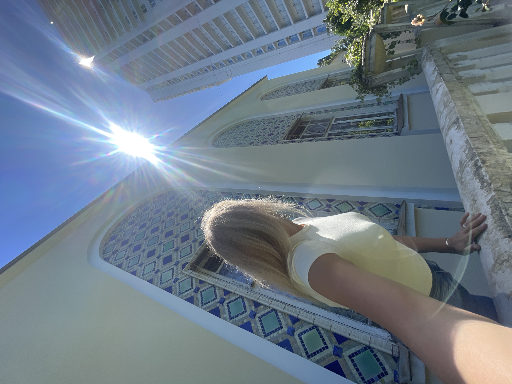 The image captures a low-angle perspective looking upwards towards a bright, sunny sky and architectural structures in Sochi, Russia. In the foreground, a woman with long, flowing blond hair, wearing a light yellow t-shirt and dark bottoms, is visible from behind and partially from the side. Her right arm is extended, with her hand resting on or gripping a light-colored, textured balustrade or wall on the right side of the frame.
To the left of the woman, a distinctive building with smooth, light-colored walls features large, arched indentations filled with intricate geometric tiles in shades of blue and green, arranged in a diamond pattern. Windows are visible within these tiled sections. Further in the background, a tall, modern high-rise building with numerous balconies stands prominently against the clear, vibrant blue sky.
A brilliant, starburst-like sun dominates the upper-middle left of the sky, creating significant lens flare and indicating a clear, bright daytime. Green foliage and vines are visible cascading over a structure on the upper right, suggesting a pleasant, warm environment. No discernible text is present in the image.