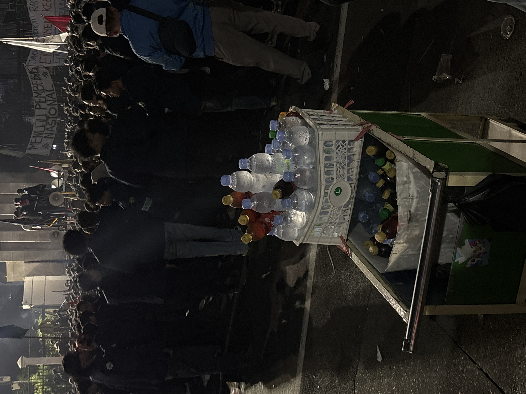 A crowd of people are gathered outdoors at night. The image is rotated 90 degrees counter-clockwise. In the foreground, a shopping cart is filled with bottled water and other drinks. The cart has a white plastic basket on top, holding clear plastic bottles with blue caps, and some bottles with yellow and red caps. Behind the cart, a person is wearing a blue jacket and a white cap, with a black bag slung over their shoulder. The person is looking down. The crowd extends into the background, with many people visible, some holding banners. One banner has text in Indonesian: "MENUJU PEMBANGUNAN NASIONAL". Other banners have red and white flags. The overall atmosphere appears to be a gathering or protest, given the banners and the large crowd. The lighting suggests it is nighttime. The location is identified as Central Jakarta, Indonesia.